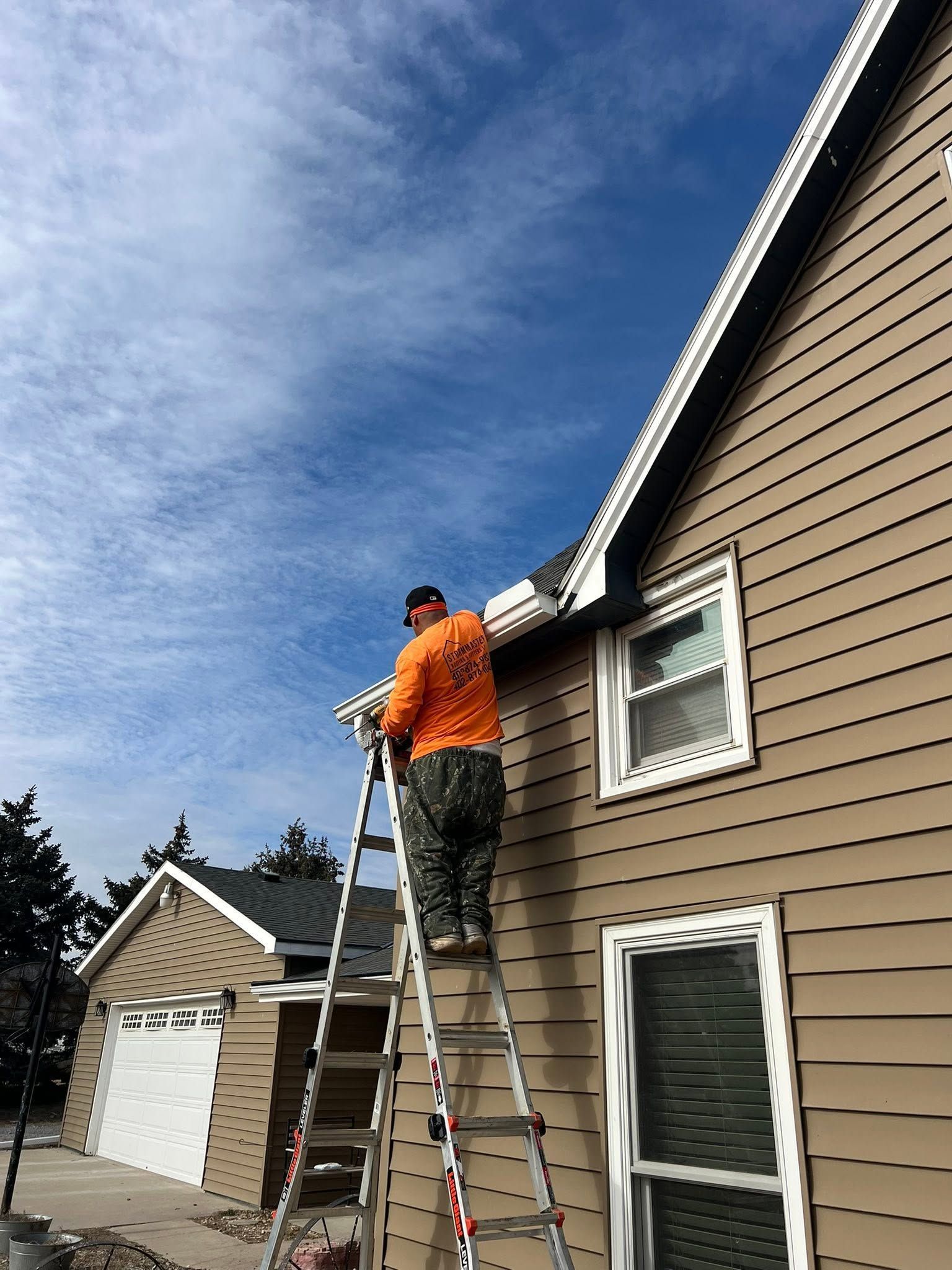 Person on a ladder painting the side of a beige house under a blue sky