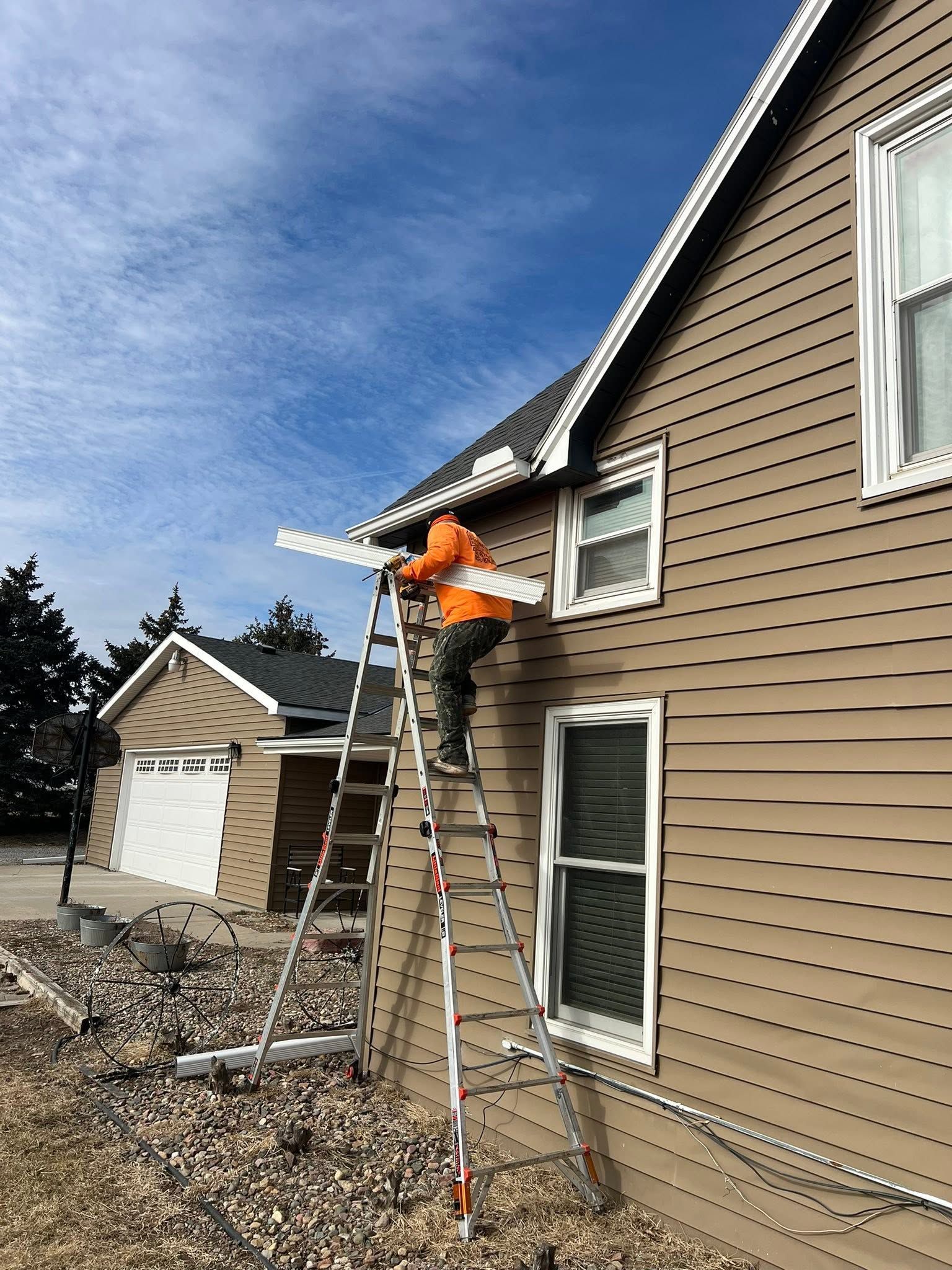 Worker on a ladder installing white trim on a beige house exterior under a blue sky.