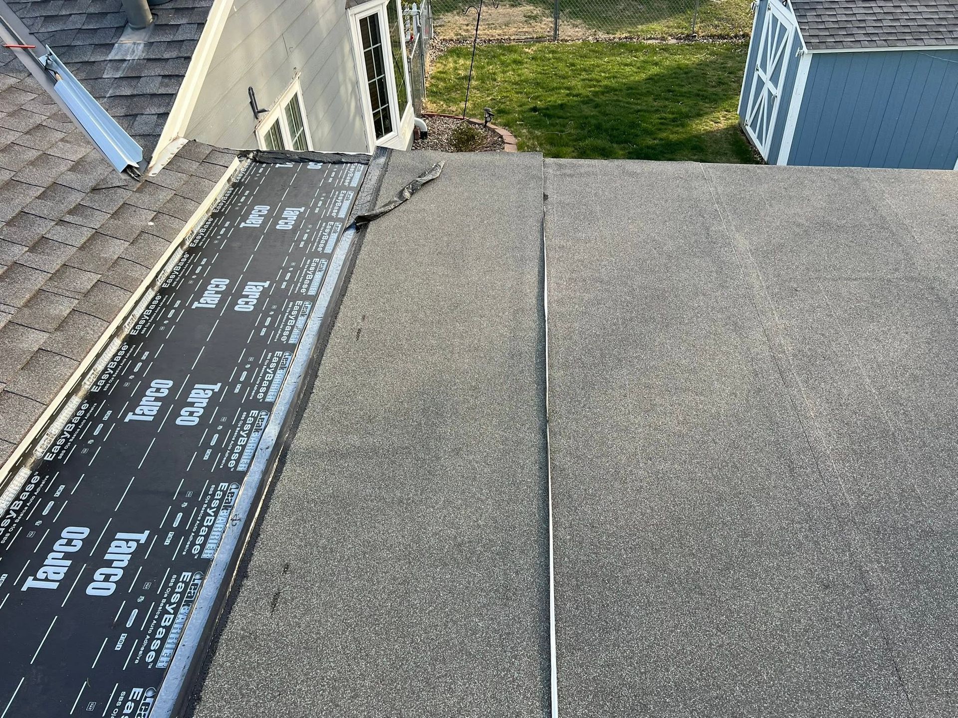 Flat rooftop with gray shingles, roof underlayment, and a seam near the edge beside neighboring houses