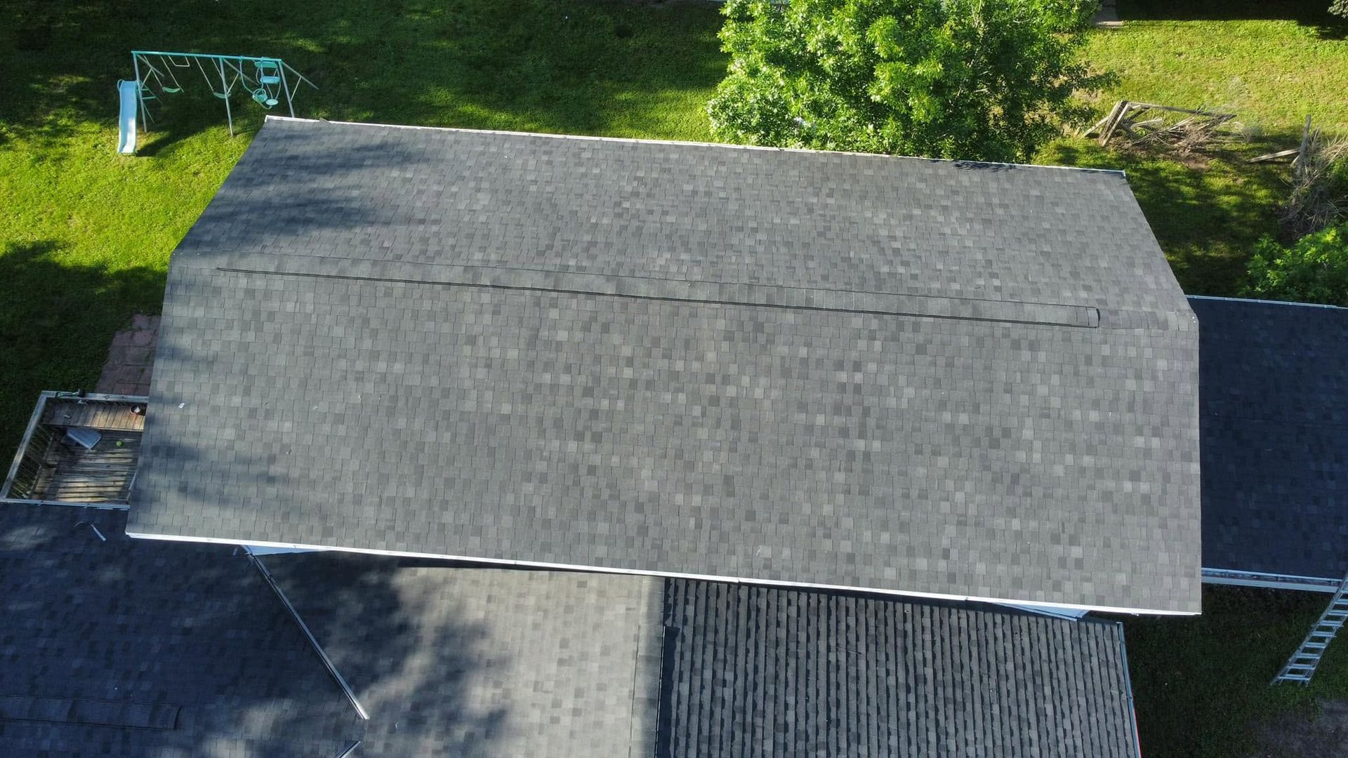 Overhead view of a gray shingled roof beside green trees and a driveway.