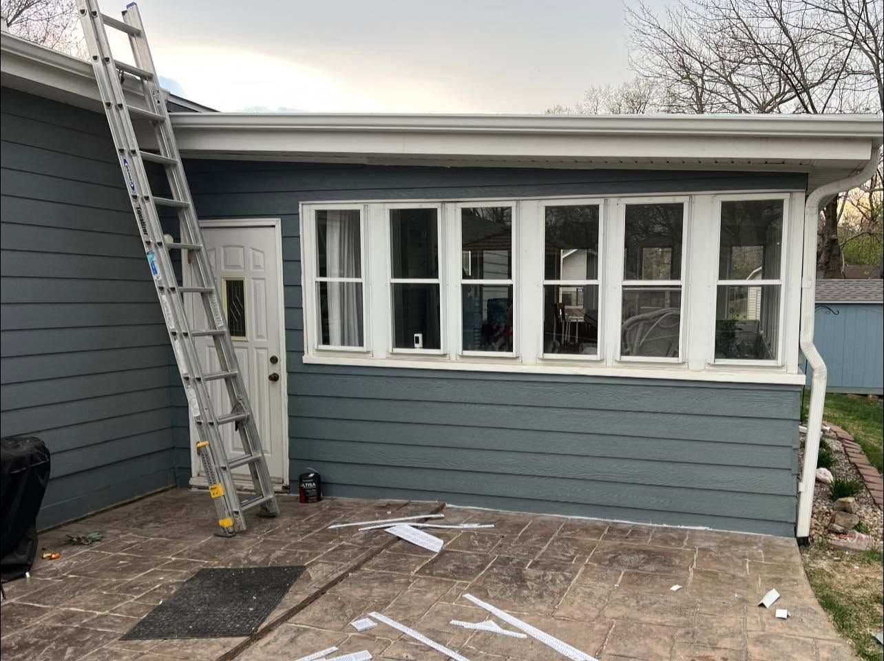 Exterior of a gray house with white-trim windows, a ladder on the left, and patio work in progress