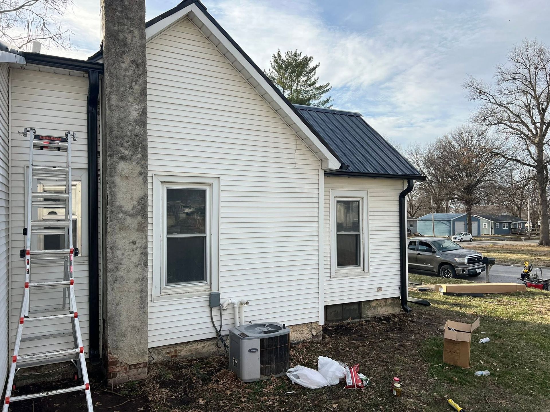 White house side with ladder against chimney, metal roof, and yard debris near a window.