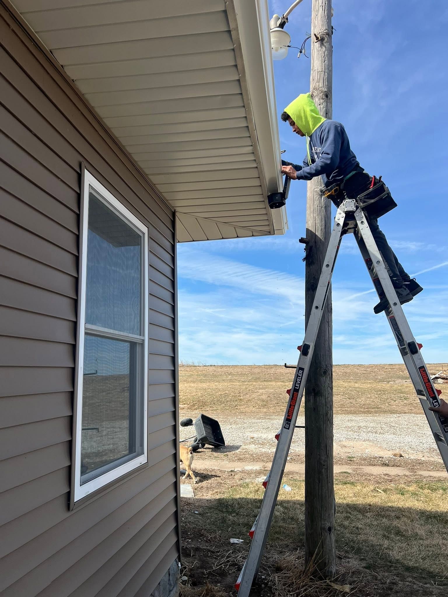 Worker on ladder painting soffit beside a house, outdoors in a rural yard.