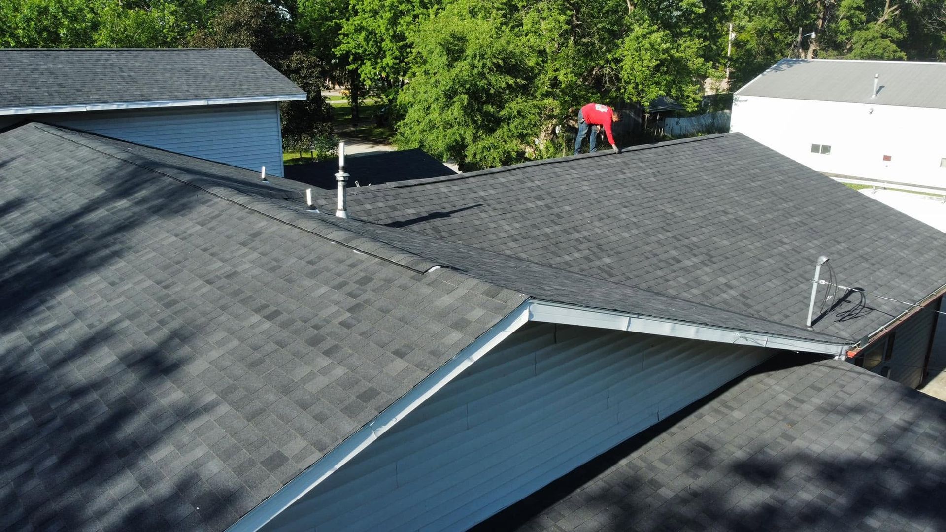 Rooftops with dark shingles and two workers inspecting a roof amid green trees