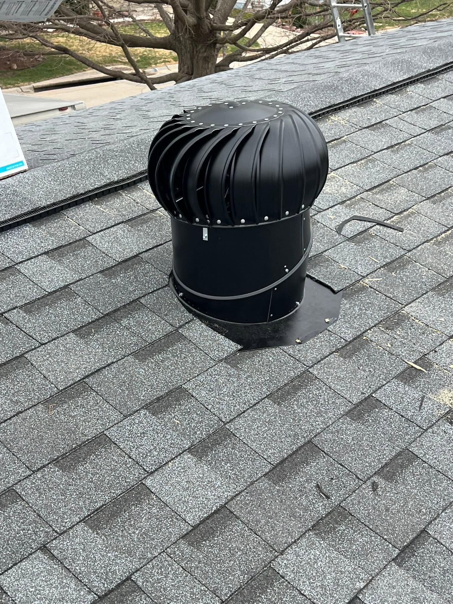 Black rooftop turbine vent on gray shingle roof near trees.