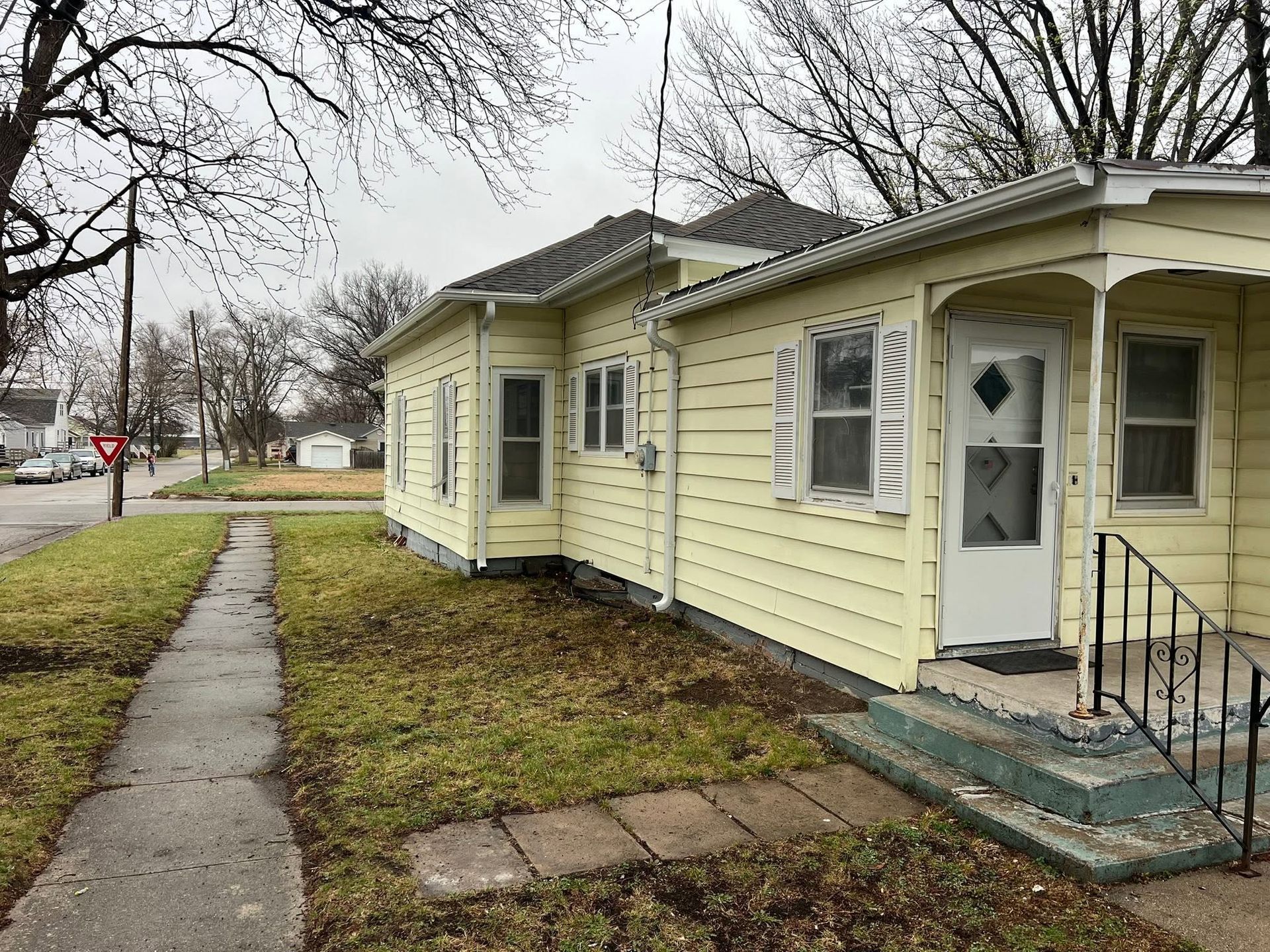 Small yellow house with white trim beside a sidewalk on a cloudy day