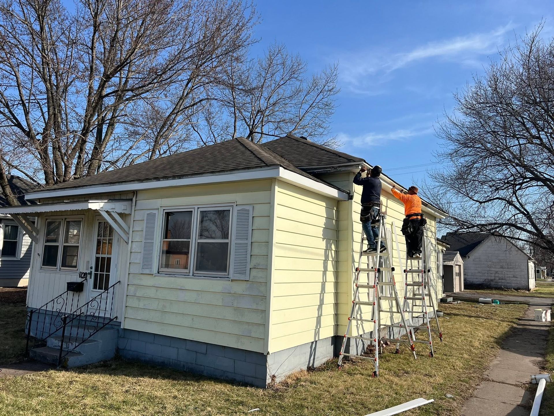 Workers on ladders repairing the roof of a small pale yellow house on a sunny day
