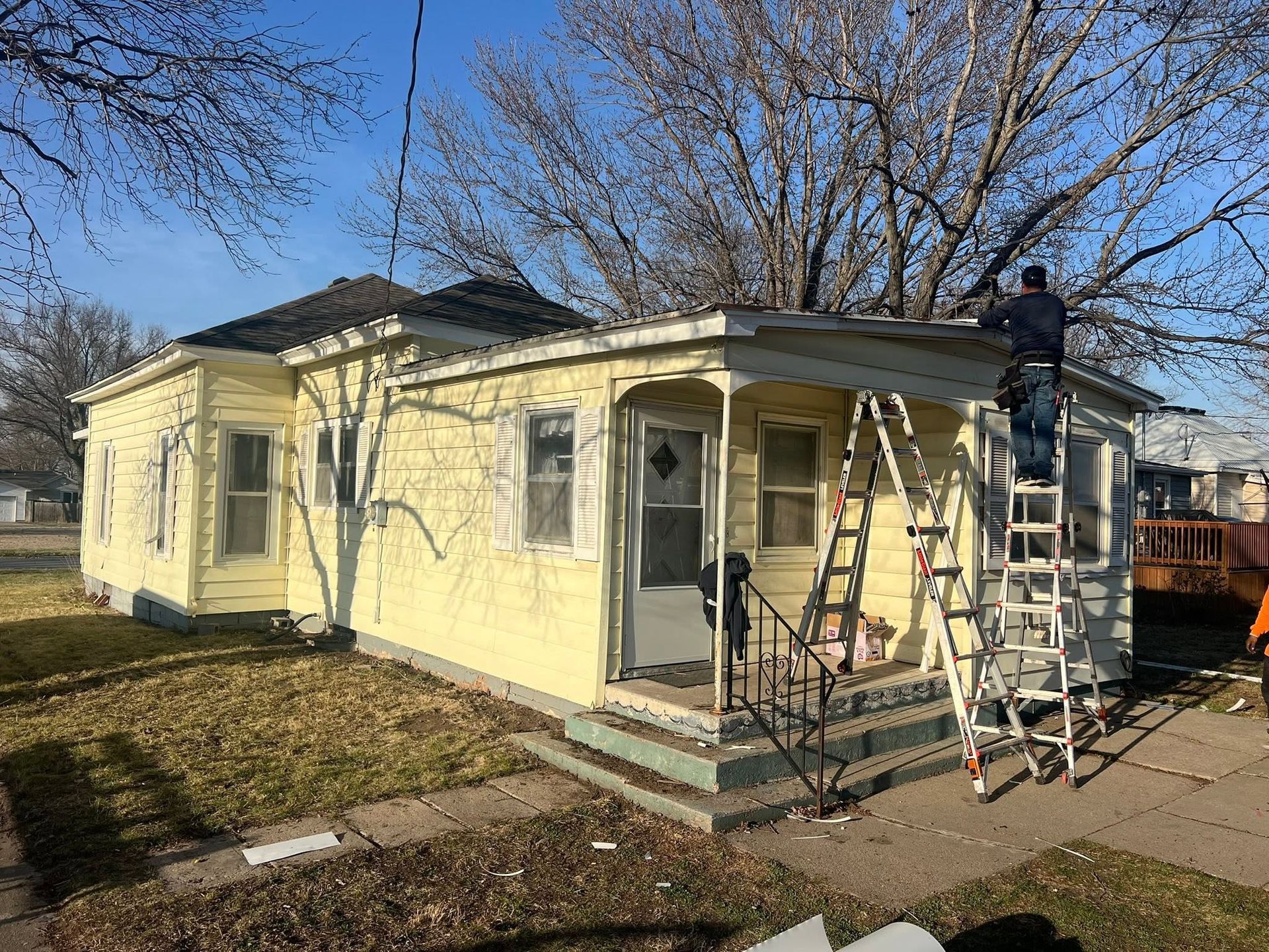 Two workers on ladders repainting a small yellow house exterior on a sunny day