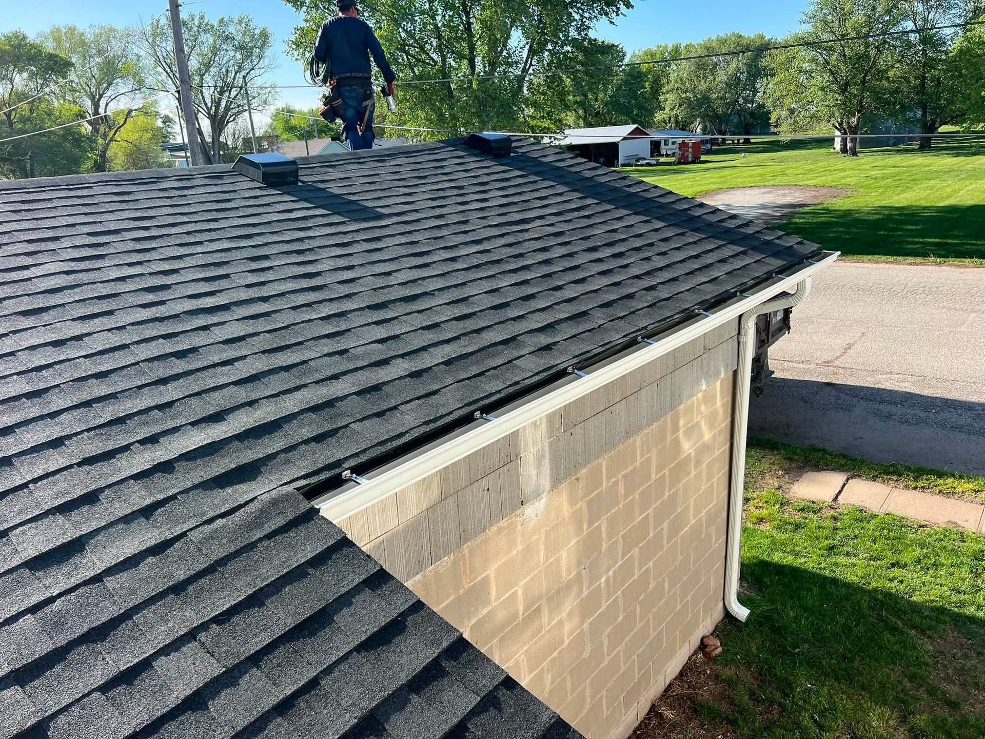 Worker on a shingled roof over a beige building beside a street and grassy yard