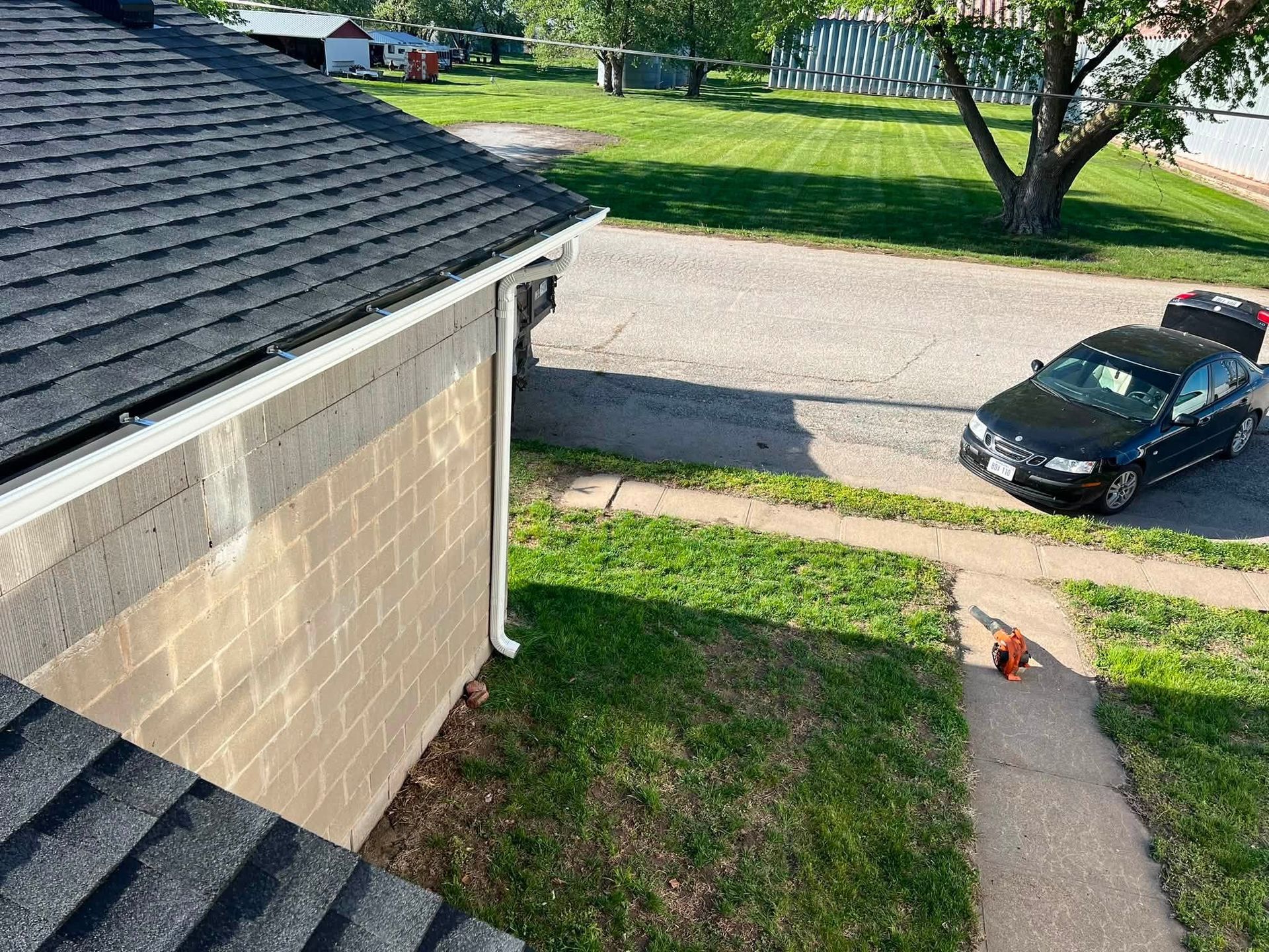 View from a roof onto a suburban street with a parked black car, driveway, lawn, and house roof shingles.