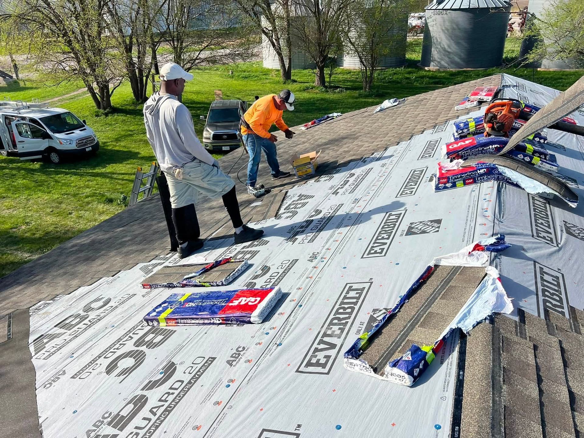 Workers installing roof shingles on a sloped roof, with materials spread out under a clear sky.
