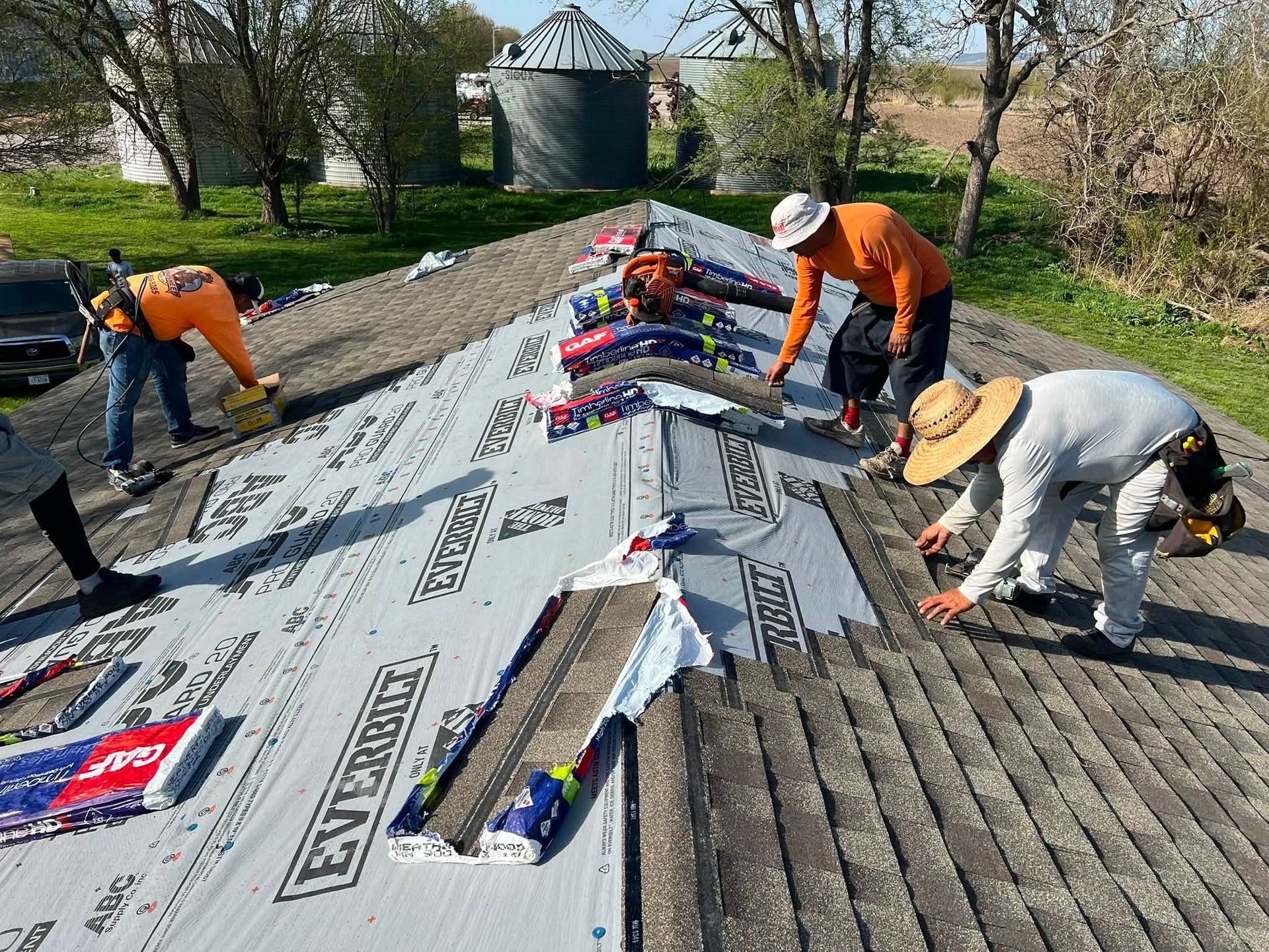 Workers installing a white membrane roof on a sloped rooftop with rolls of material and tools.