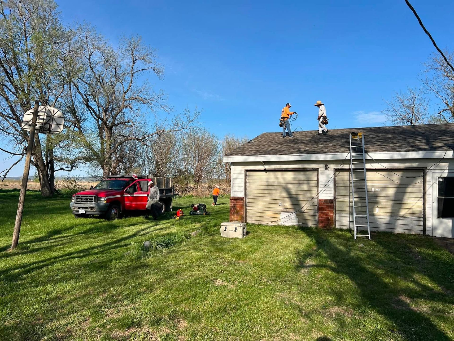 Roofers on a garage roof beside a red pickup truck in a grassy yard, under a clear blue sky.