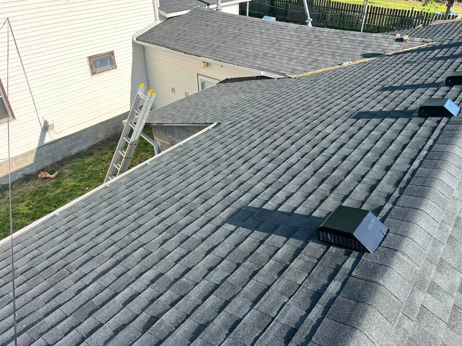 Roof with gray shingles, ladder against wall, and two roof vents in a residential backyard