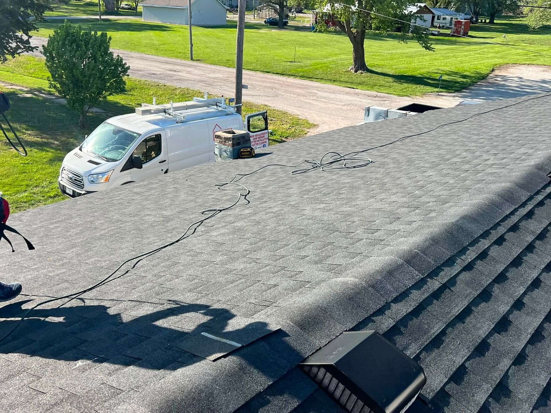 Damaged shingle roof with a white van and workers visible below on a sunny residential street