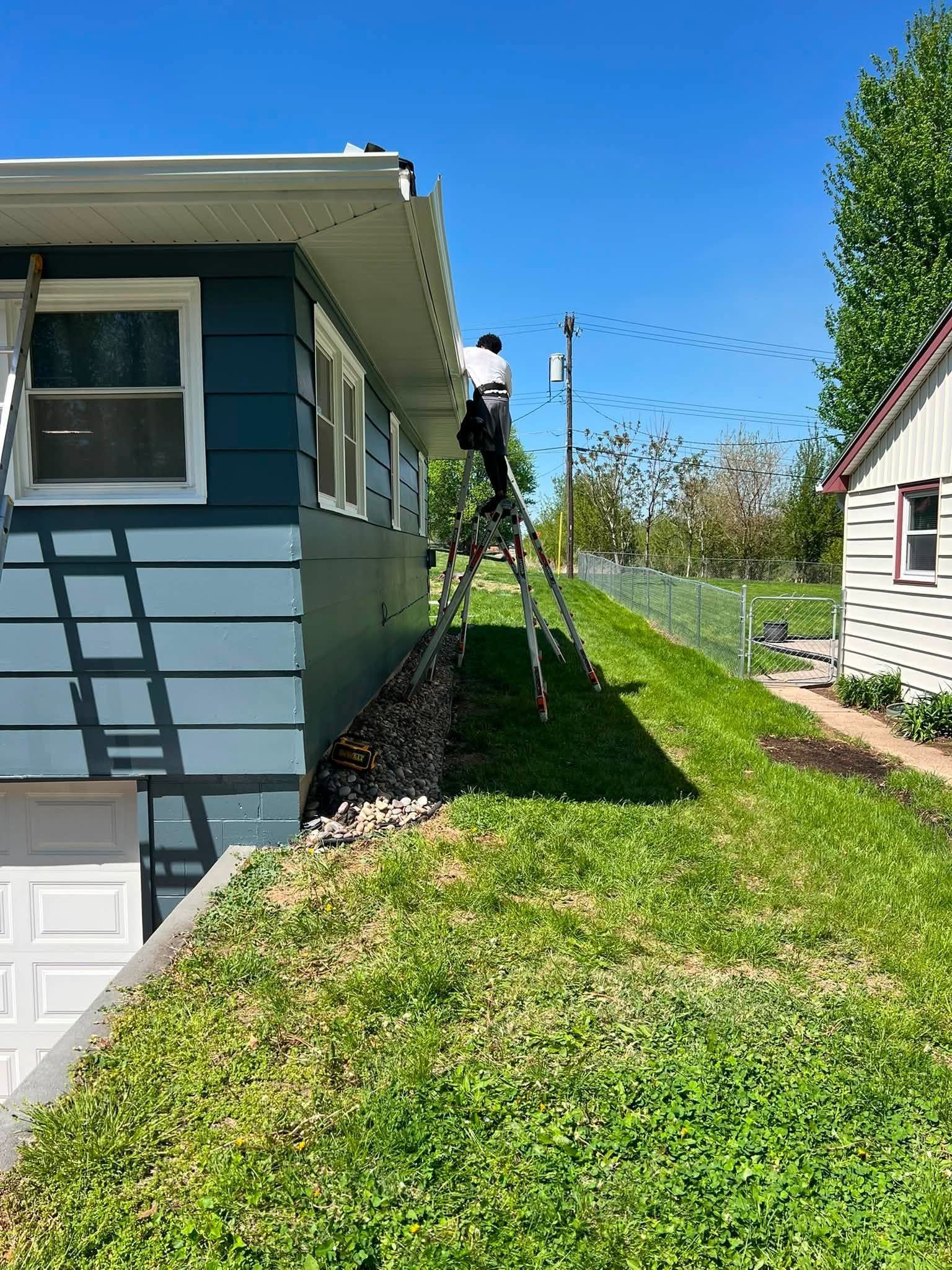 Worker on ladder beside blue house on a grassy slope under a clear sky