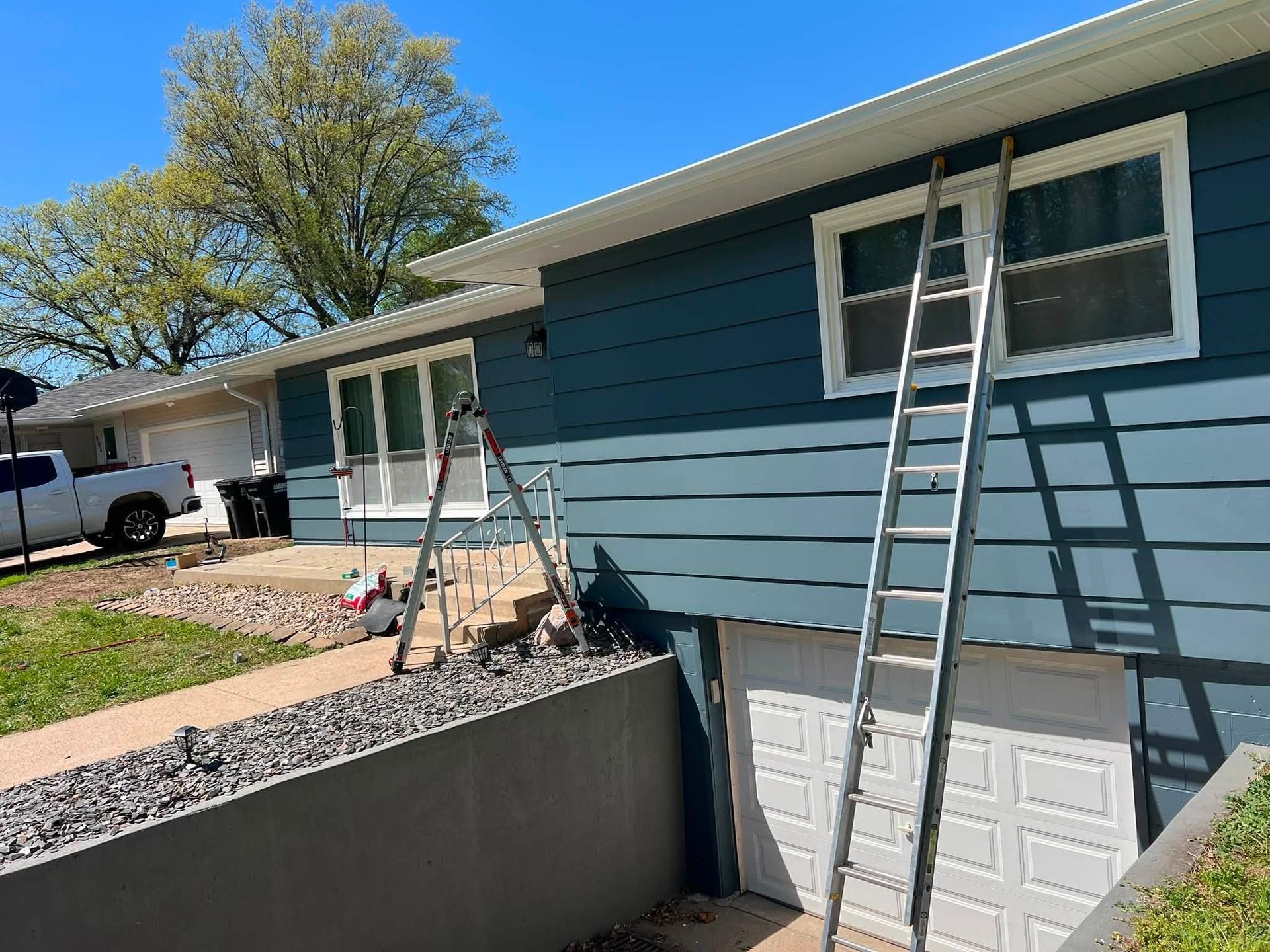 Ladders leaning against a blue house exterior with white garage doors and a raised planter wall