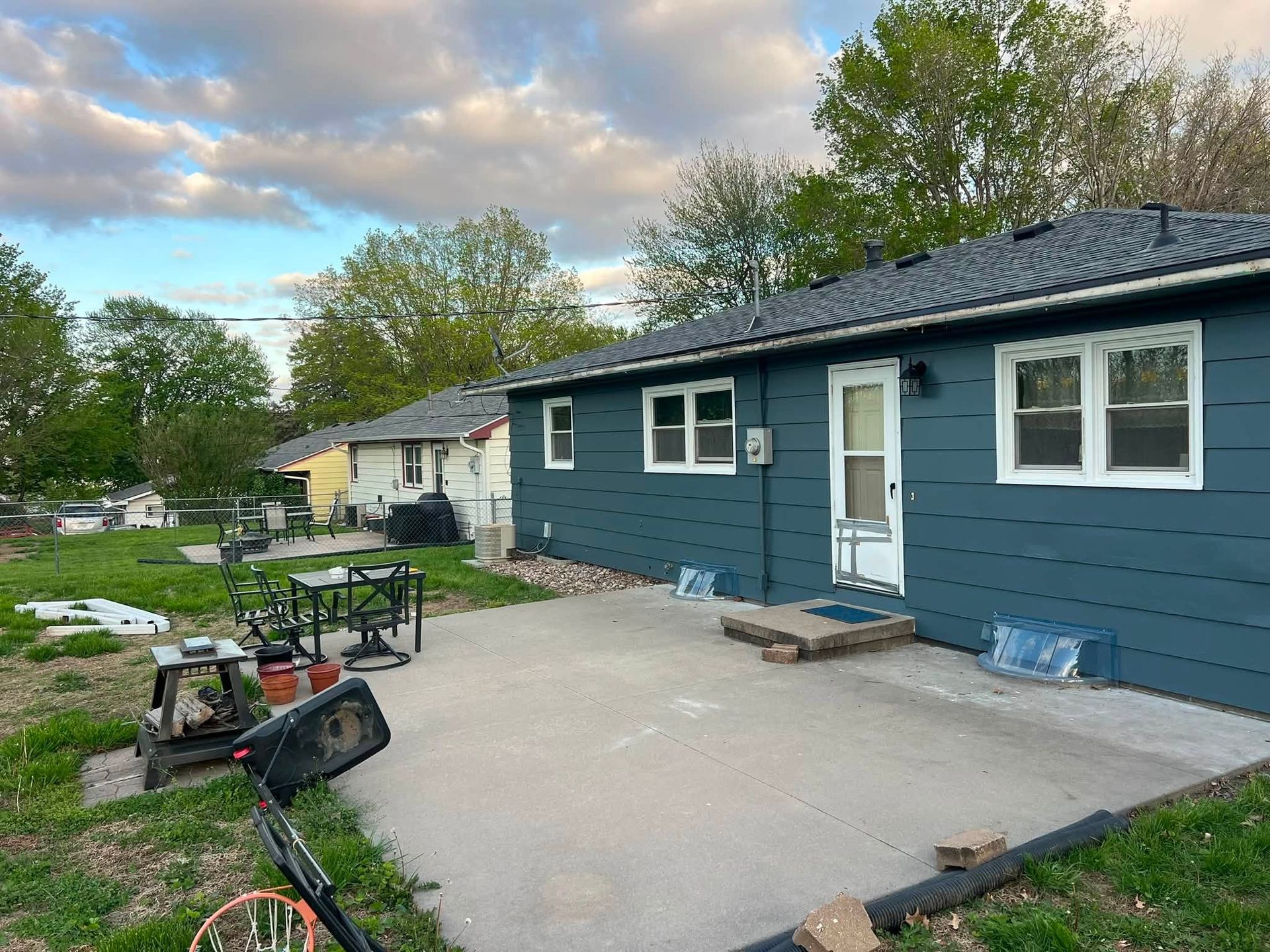 Backyard patio beside a blue house with concrete slab, lawn, and garden tools under cloudy sky