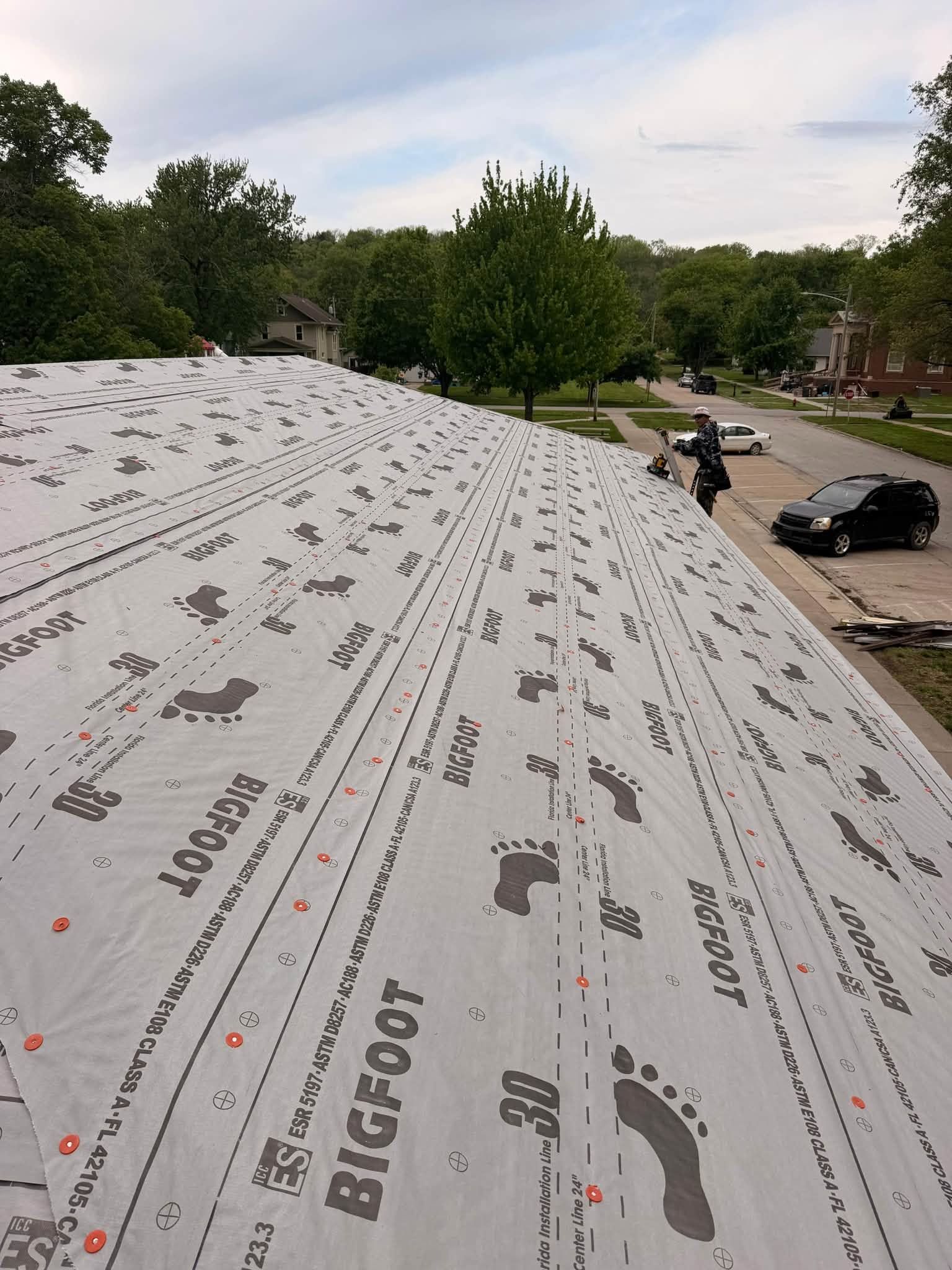 Workers installing gray roofing underlayment on a steep roof, with houses and trees below.