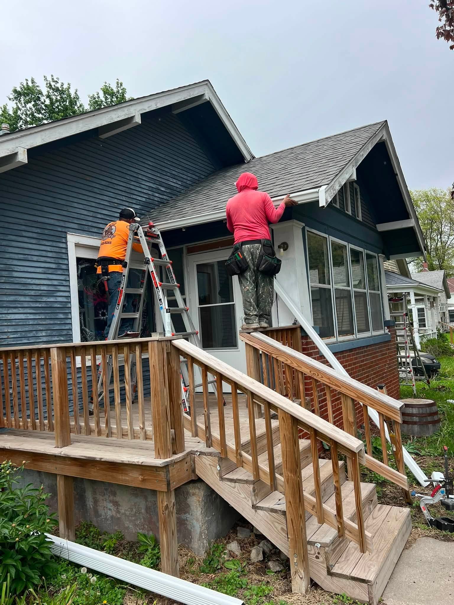 Two workers painting a dark house exterior beside a wooden porch and stairs