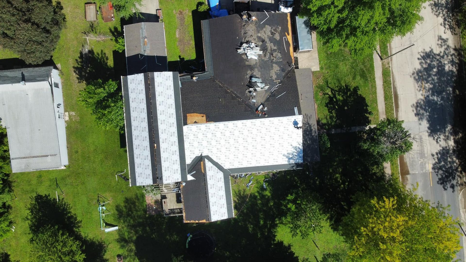 Aerial view of a house with a damaged dark roof, surrounded by trees and a road.