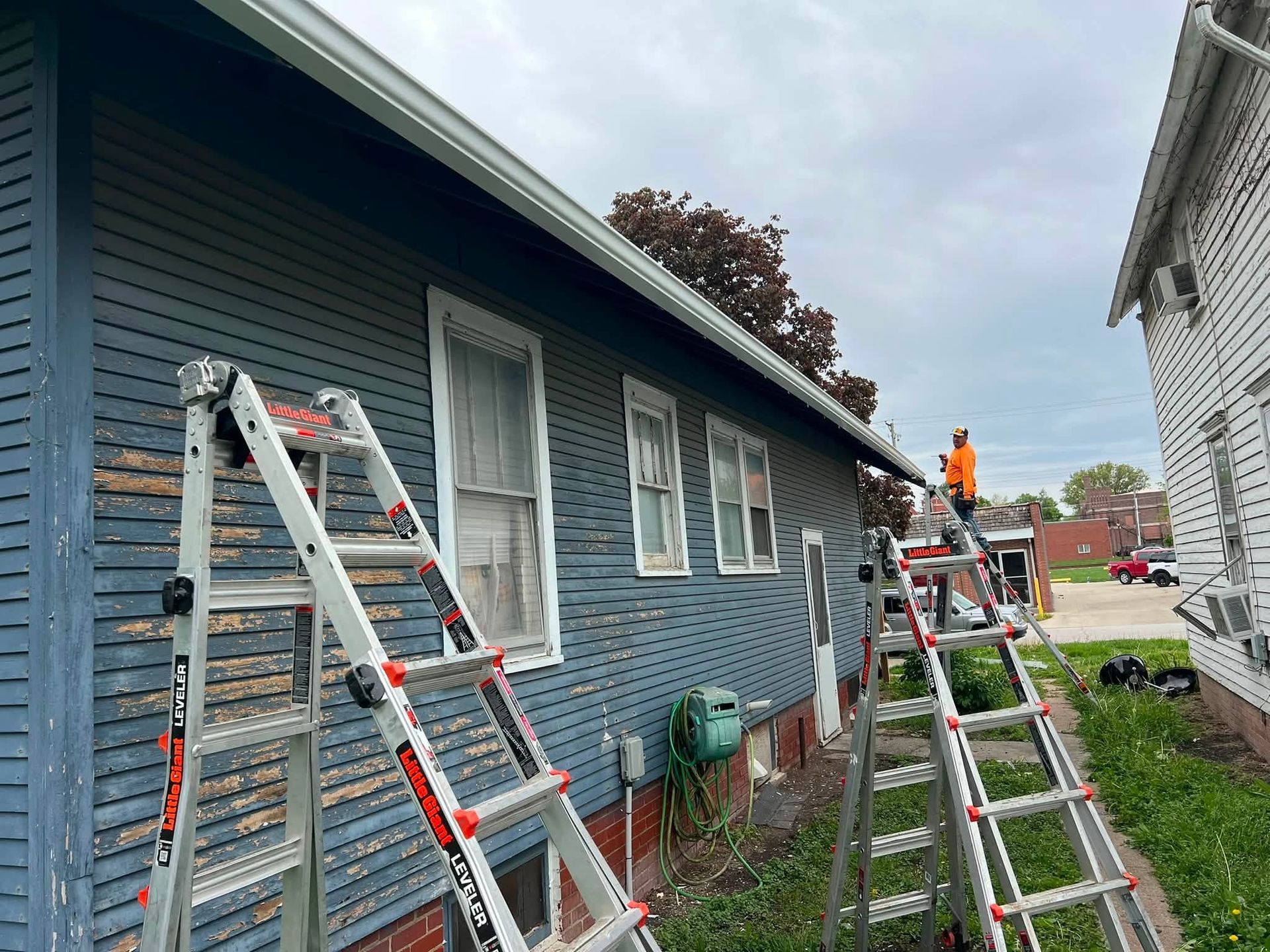 Ladders leaned against a blue house while workers do roof or gutter repairs on a cloudy day.