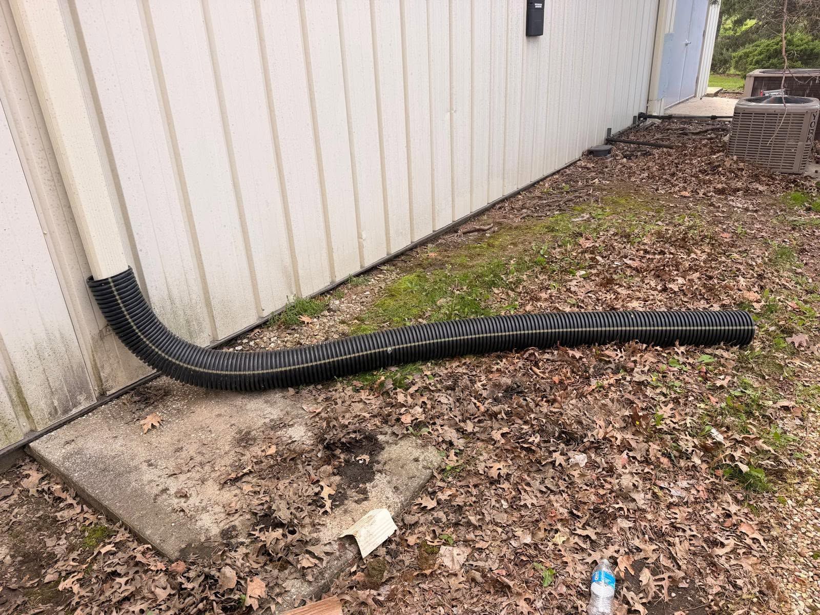 Black corrugated drain pipe extends from a white building onto muddy ground with leaves and debris.
