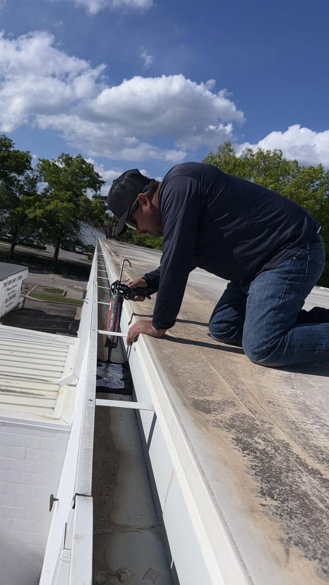 Worker crouched on a roof inspecting or repairing a white gutter under a blue sky.