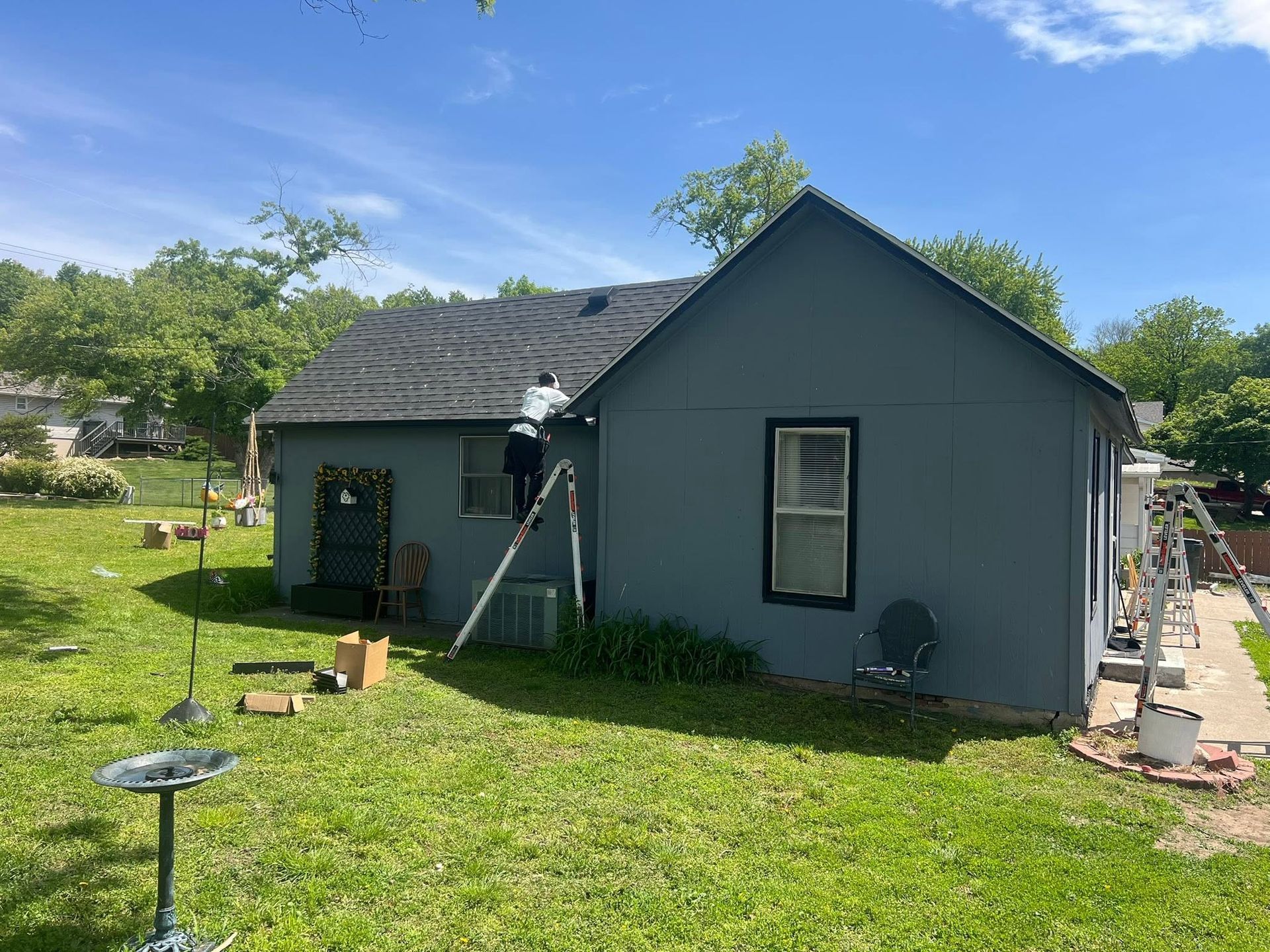 Small gray house in a grassy yard with a ladder against the roof and tools nearby.