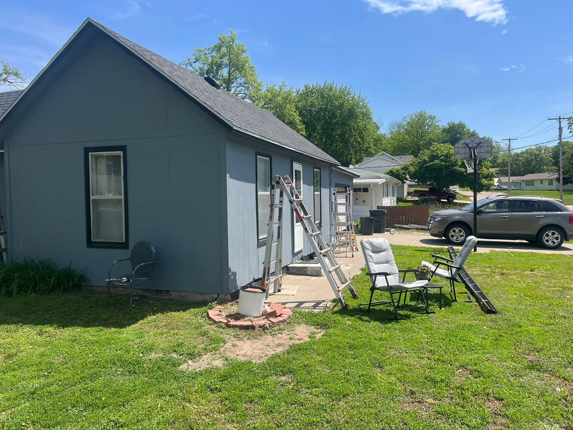 Small gray house with a ladder by the side, a chair in the yard, and a parked SUV nearby.