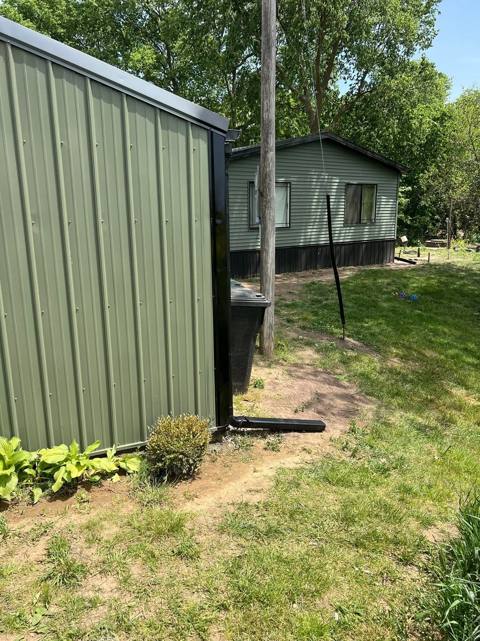 Green shed beside grassy yard with a small cabin and trees in the background