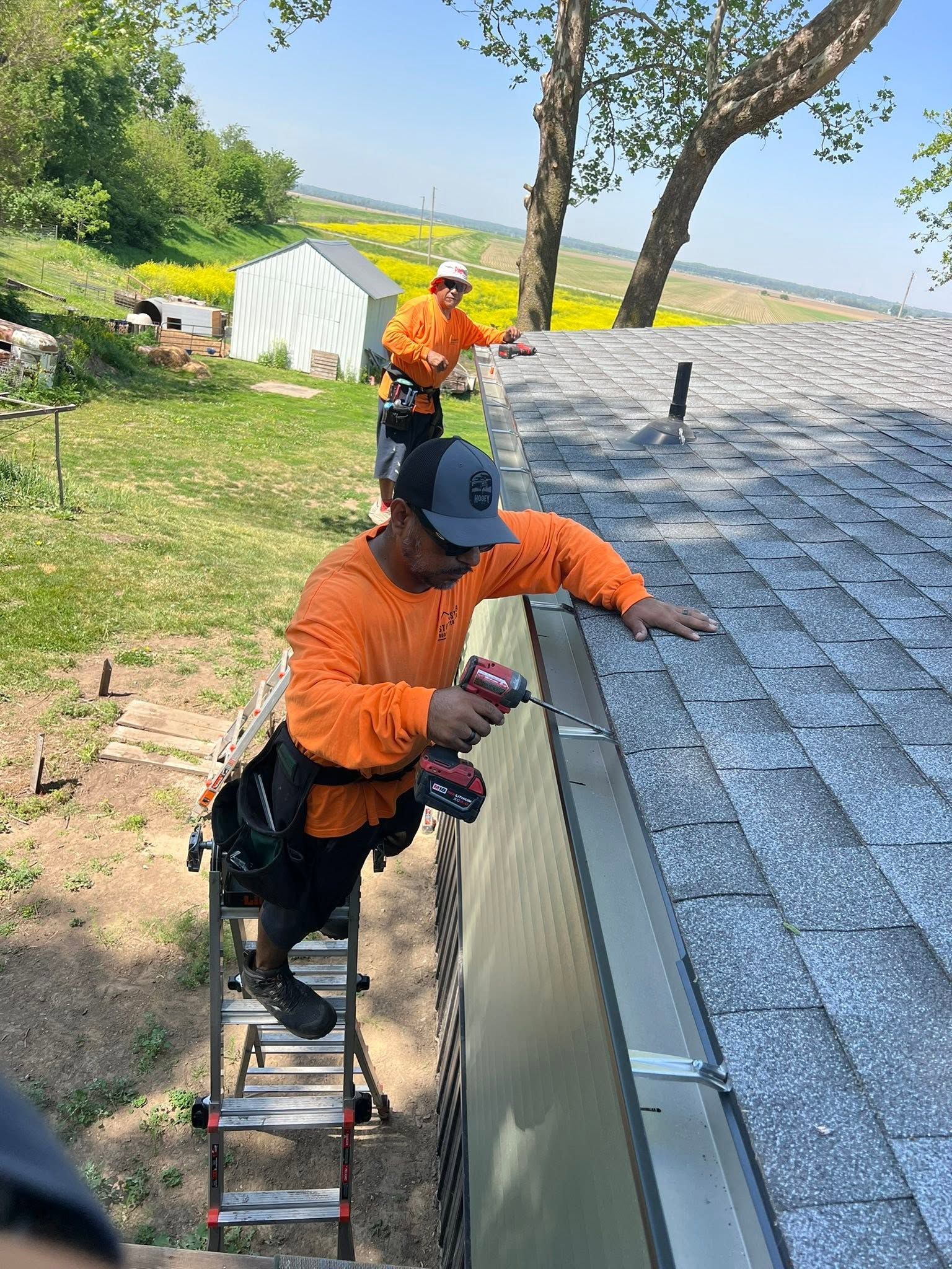 Two workers in orange shirts install roof shingles on a house near a ladder.