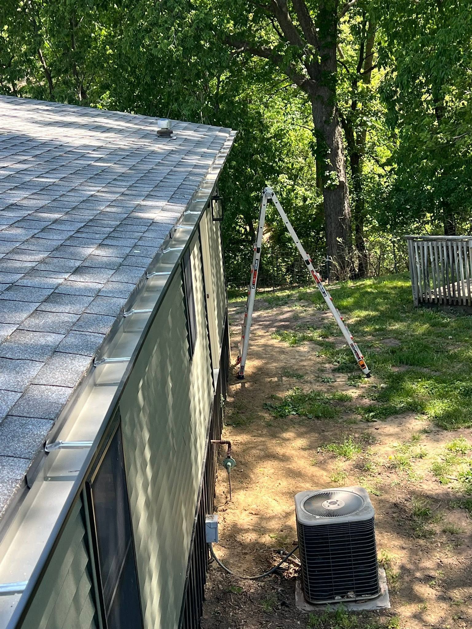 Side of a house with a ladder in a leafy yard, and an outdoor HVAC unit in the foreground.