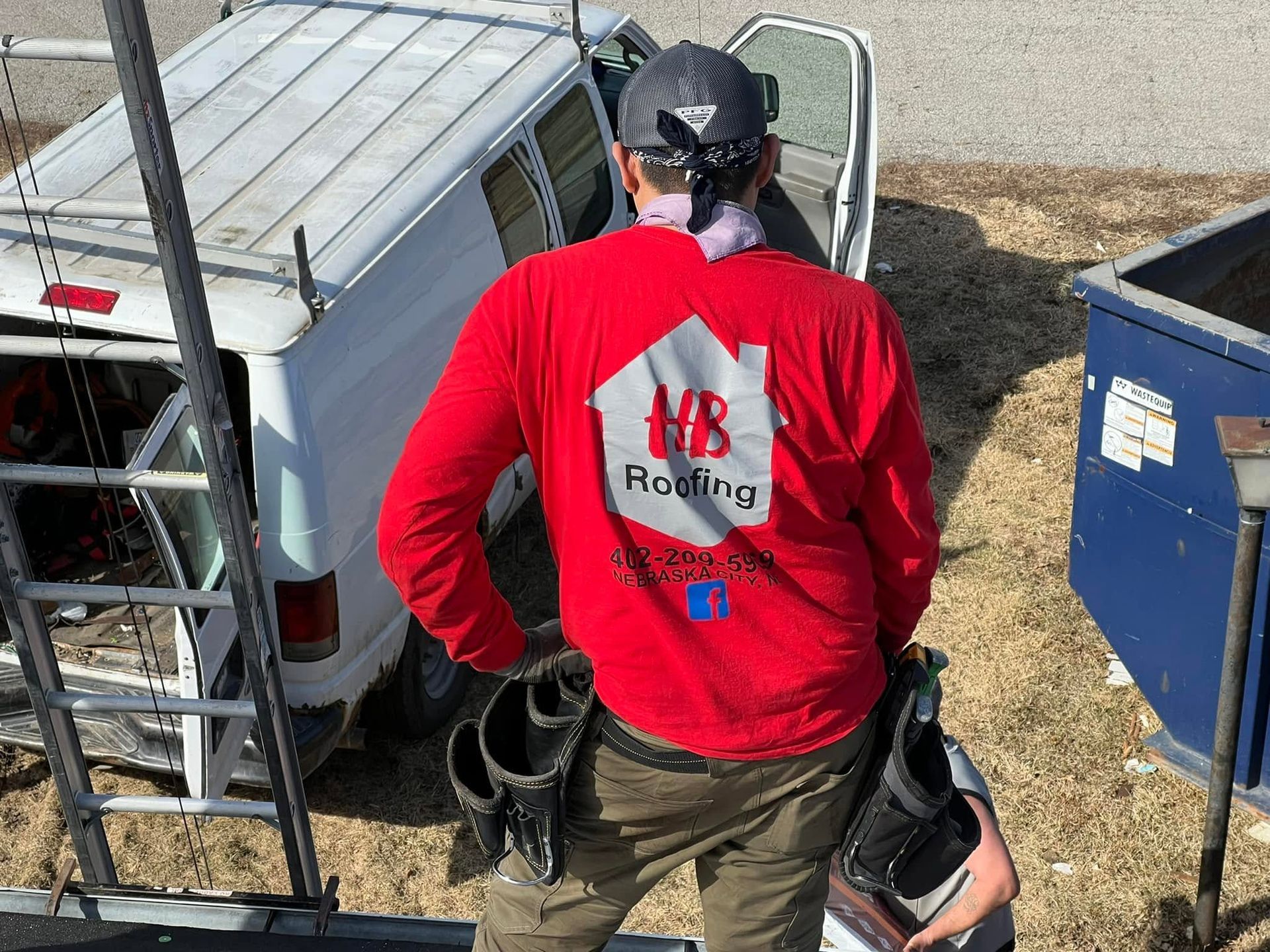 Person in red shirt carrying equipment beside a white utility van and blue dumpster outdoors