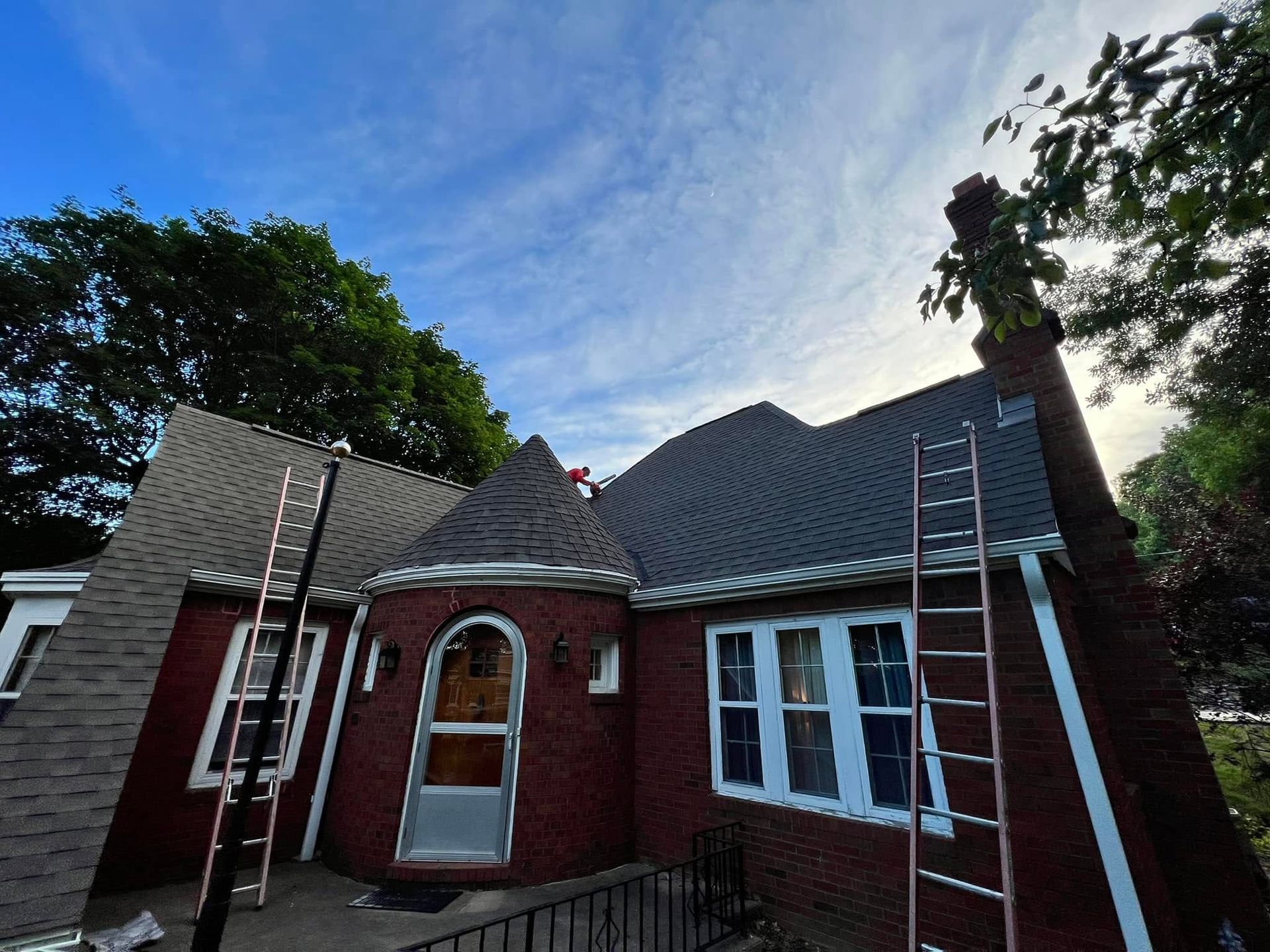 Red brick house with a steep dark roof and two ladders leaning against it under a blue sky.