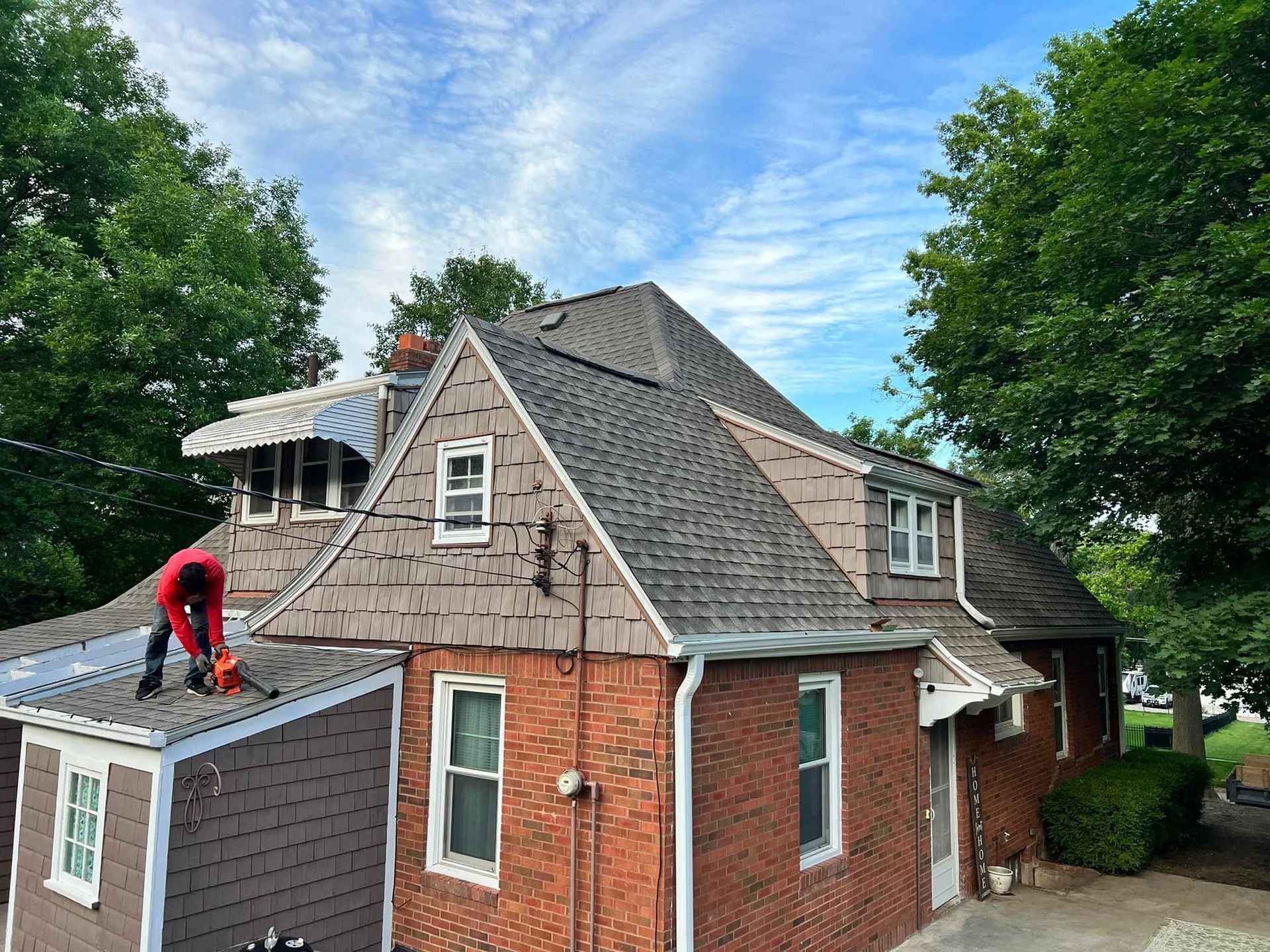 Worker on a roof repairing shingles on a brick house surrounded by trees.