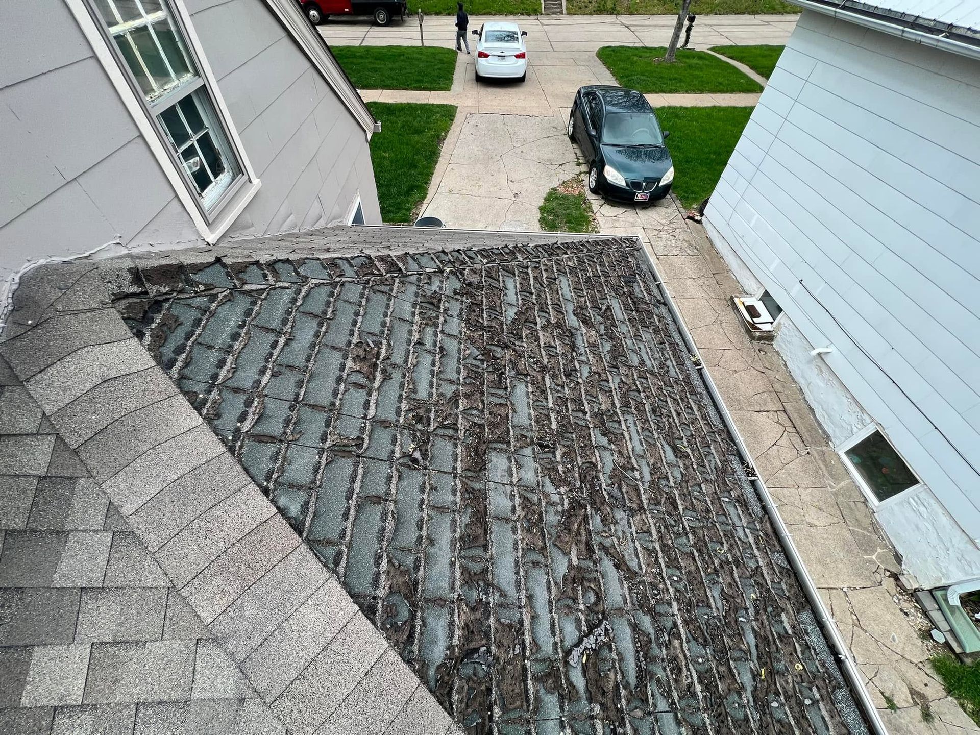 Damaged shingled roof seen from above between two houses, with driveway and parked cars below