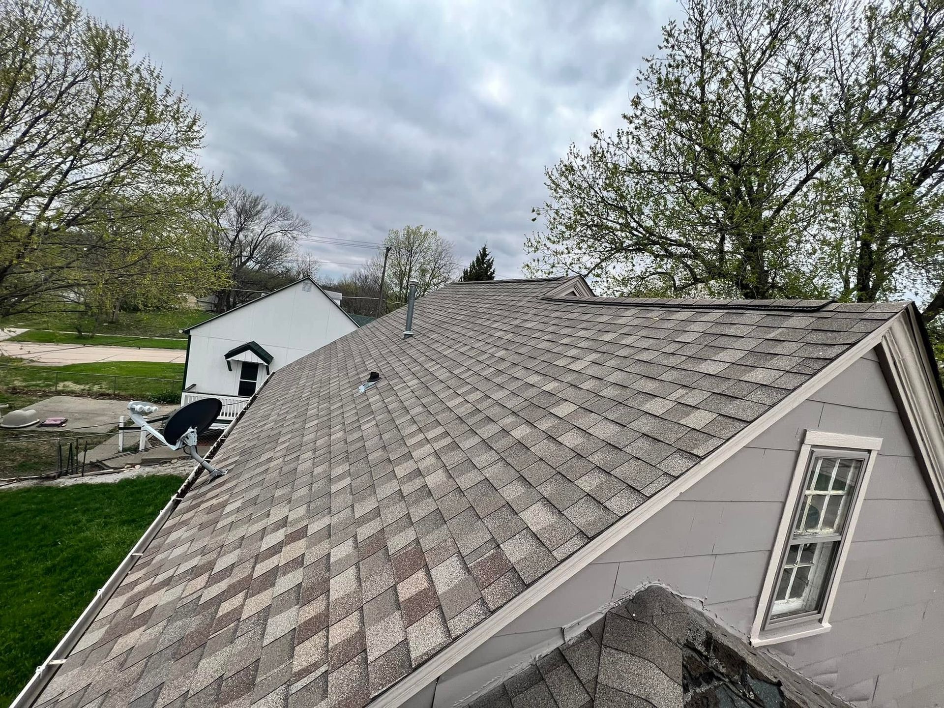 Gray shingled roof with dormer window beside a backyard and trees under cloudy sky