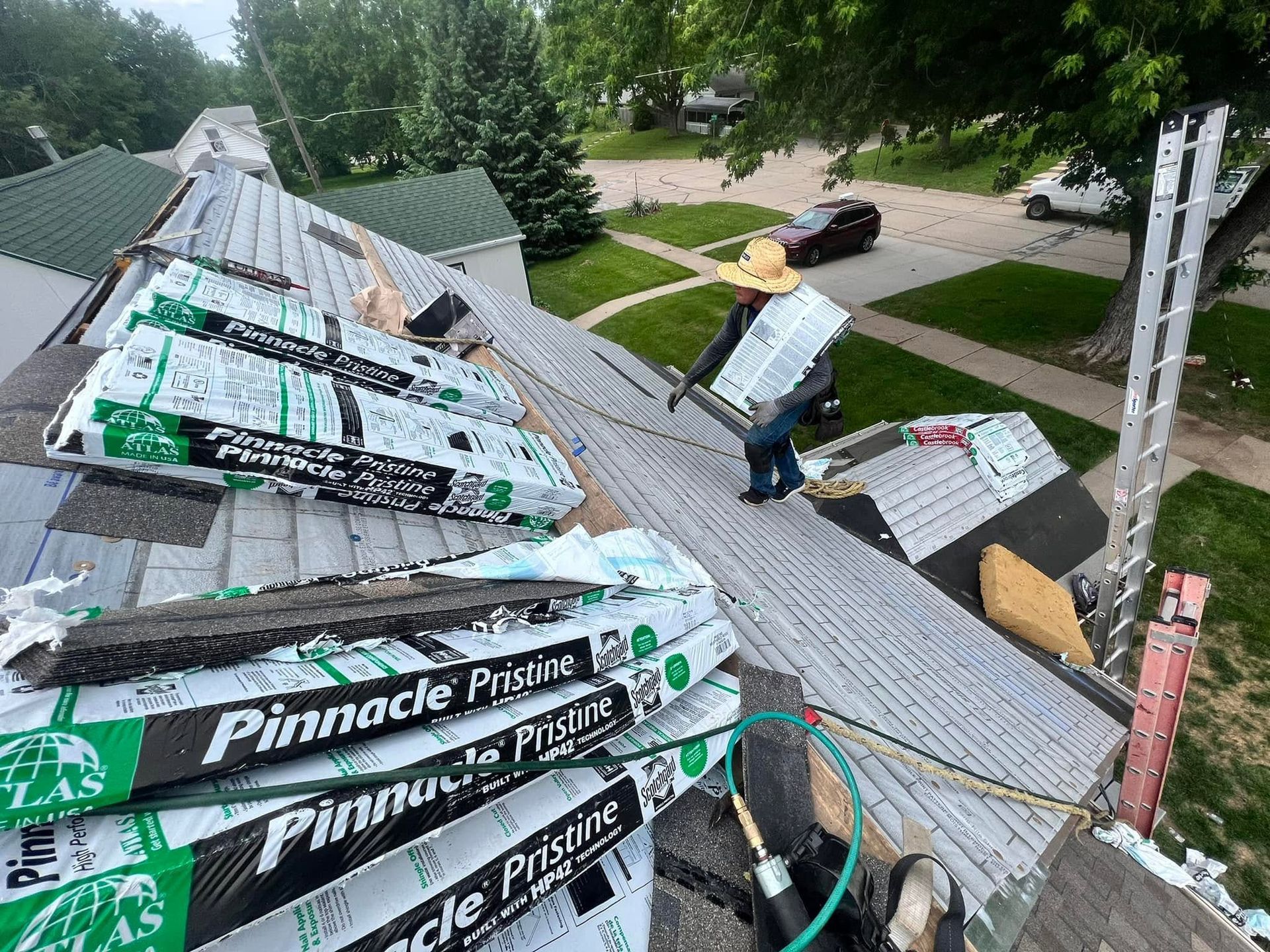 Roofers installing shingles on a steep roof beside a ladder and trees