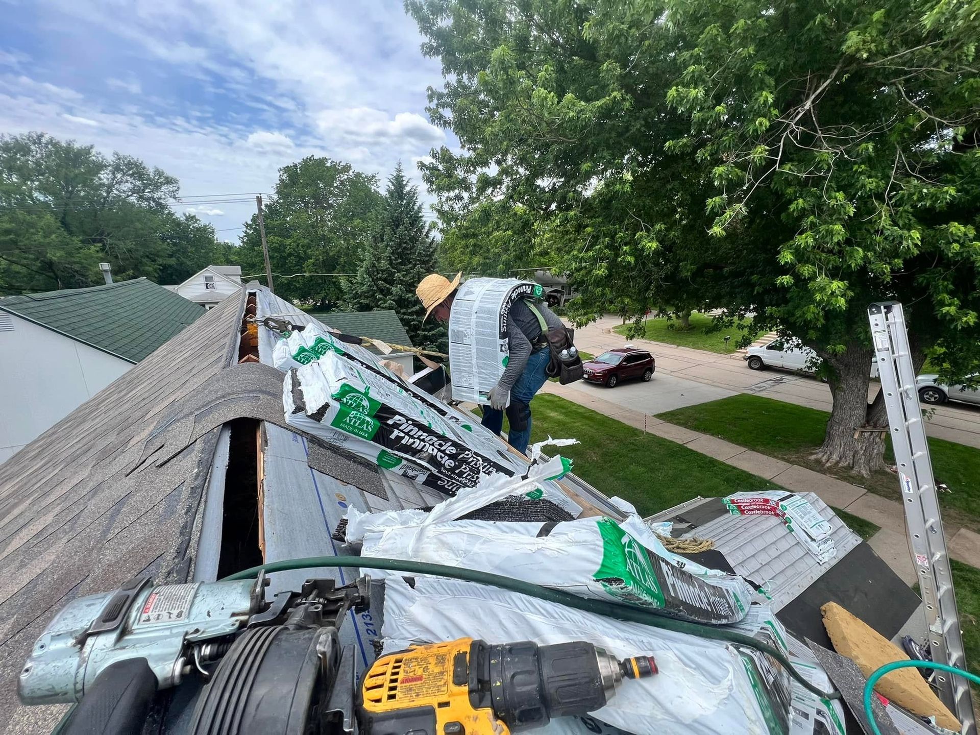 Worker replacing shingles on a steep roof with tools and roofing materials visible, trees and street below
