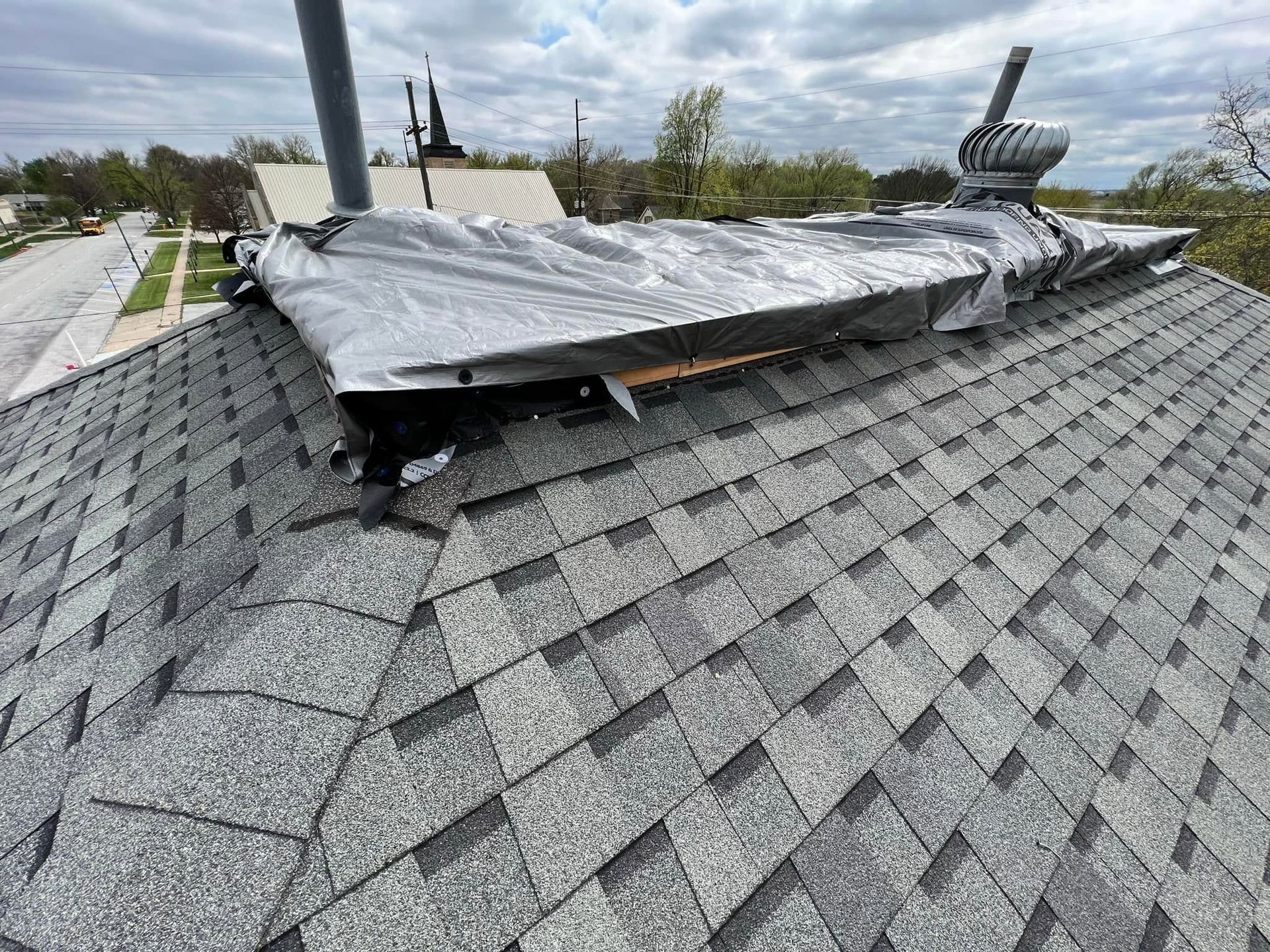 Damaged roof with a large torn gray tarp lifted over shingles in a windy outdoor setting