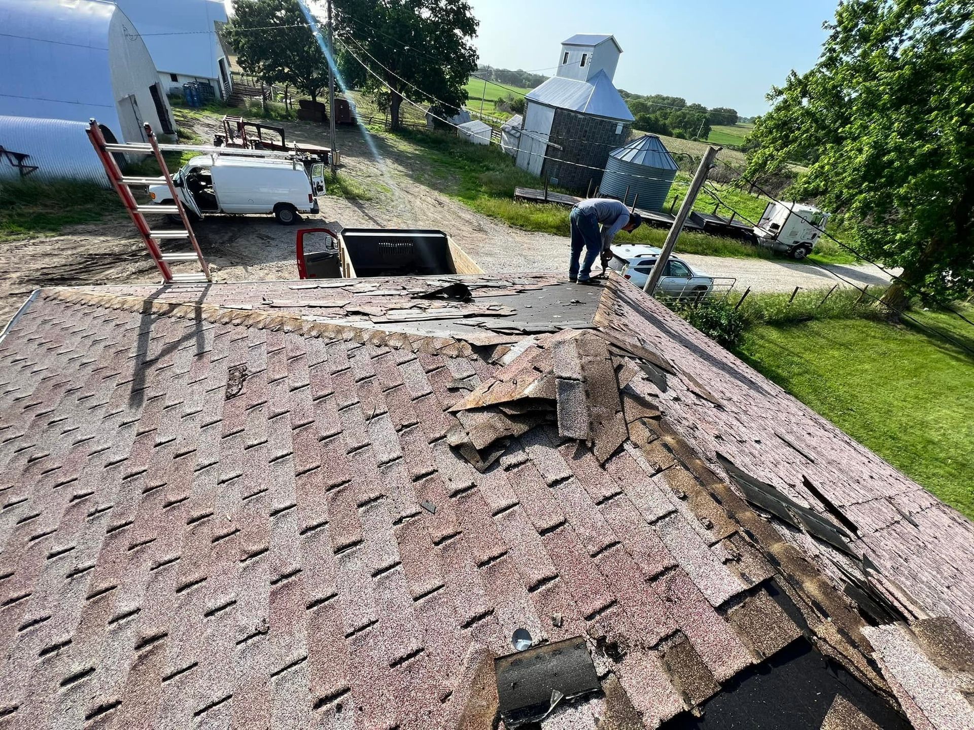 Roof shingles being repaired by a worker beside a ladder, with a white truck and rural buildings in the background