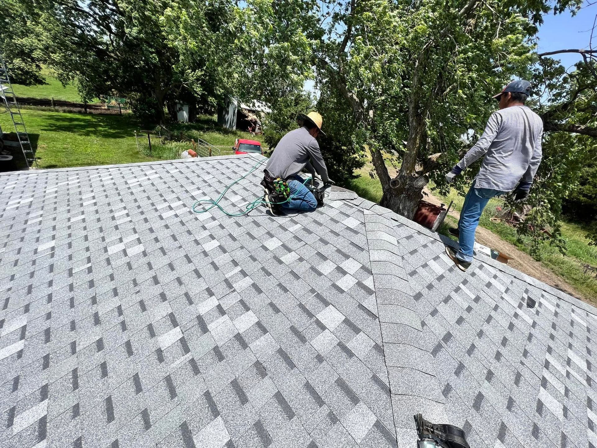 Two workers installing gray roof shingles on a sloped roof beside a tree