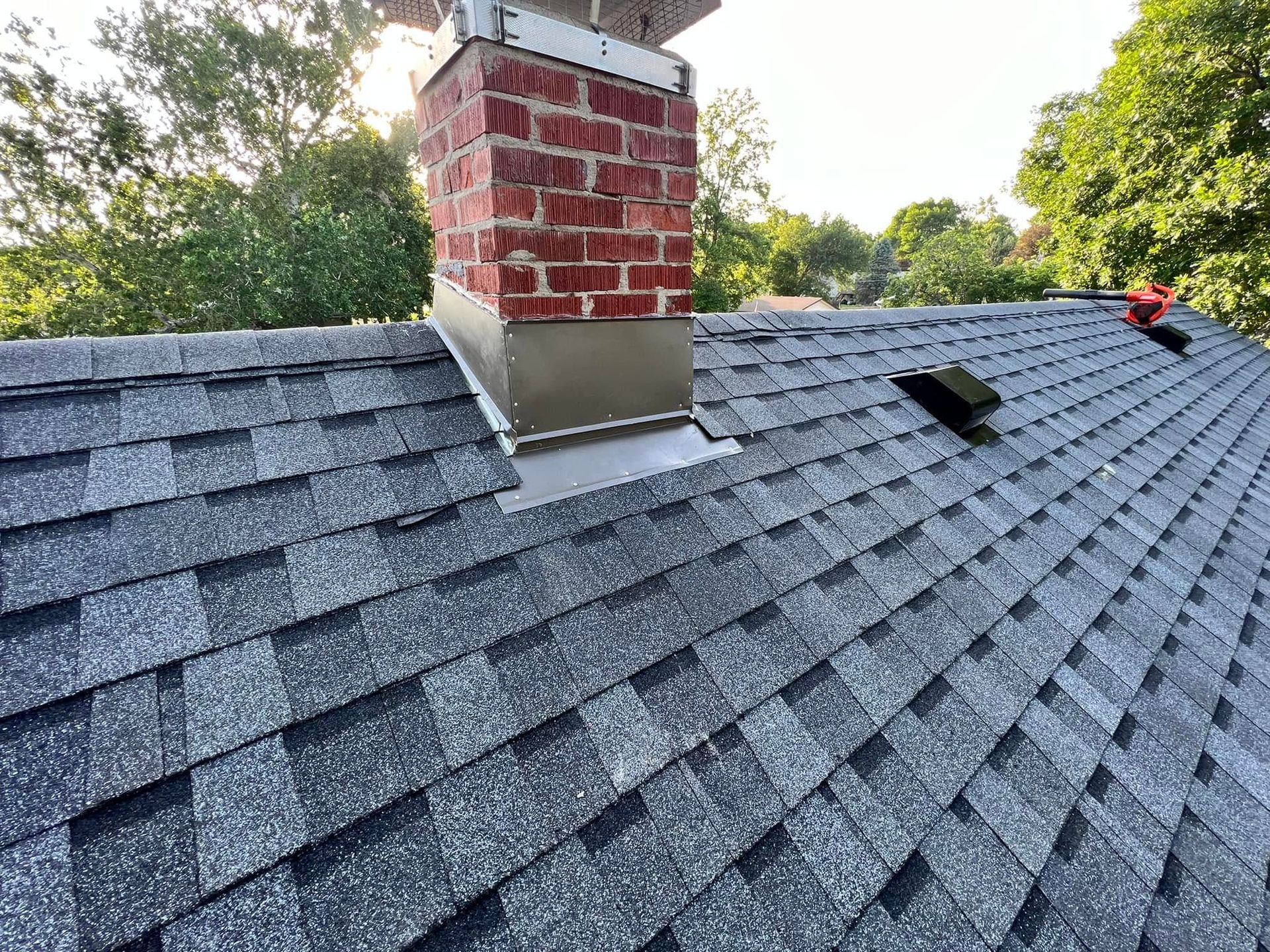 Shingled roof with red brick chimney and a small black roof vent on a sunny day