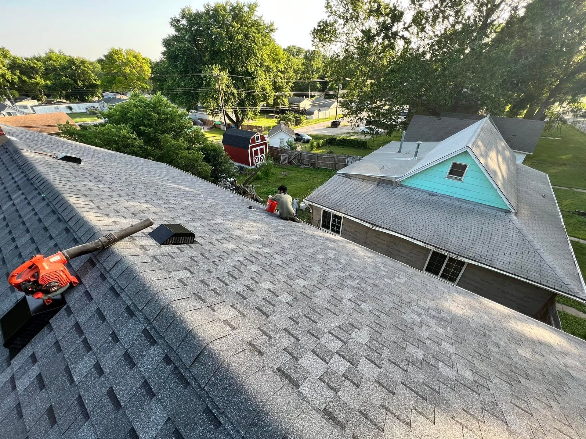 Rooftop view of gray shingles beside a turquoise house and green trees in a sunny neighborhood