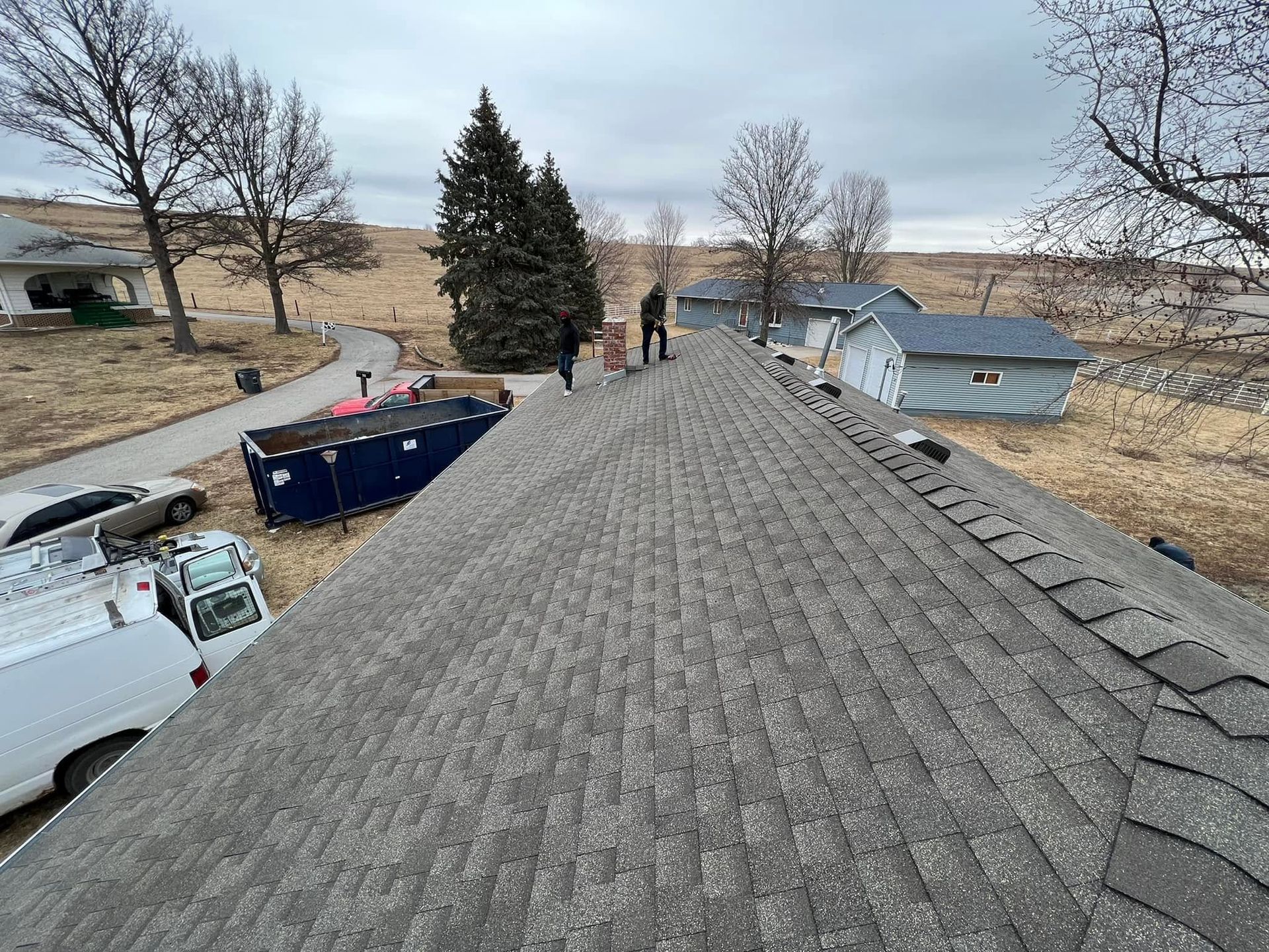 Roofline view over a rural yard with trees, trucks, and a small house in the distance