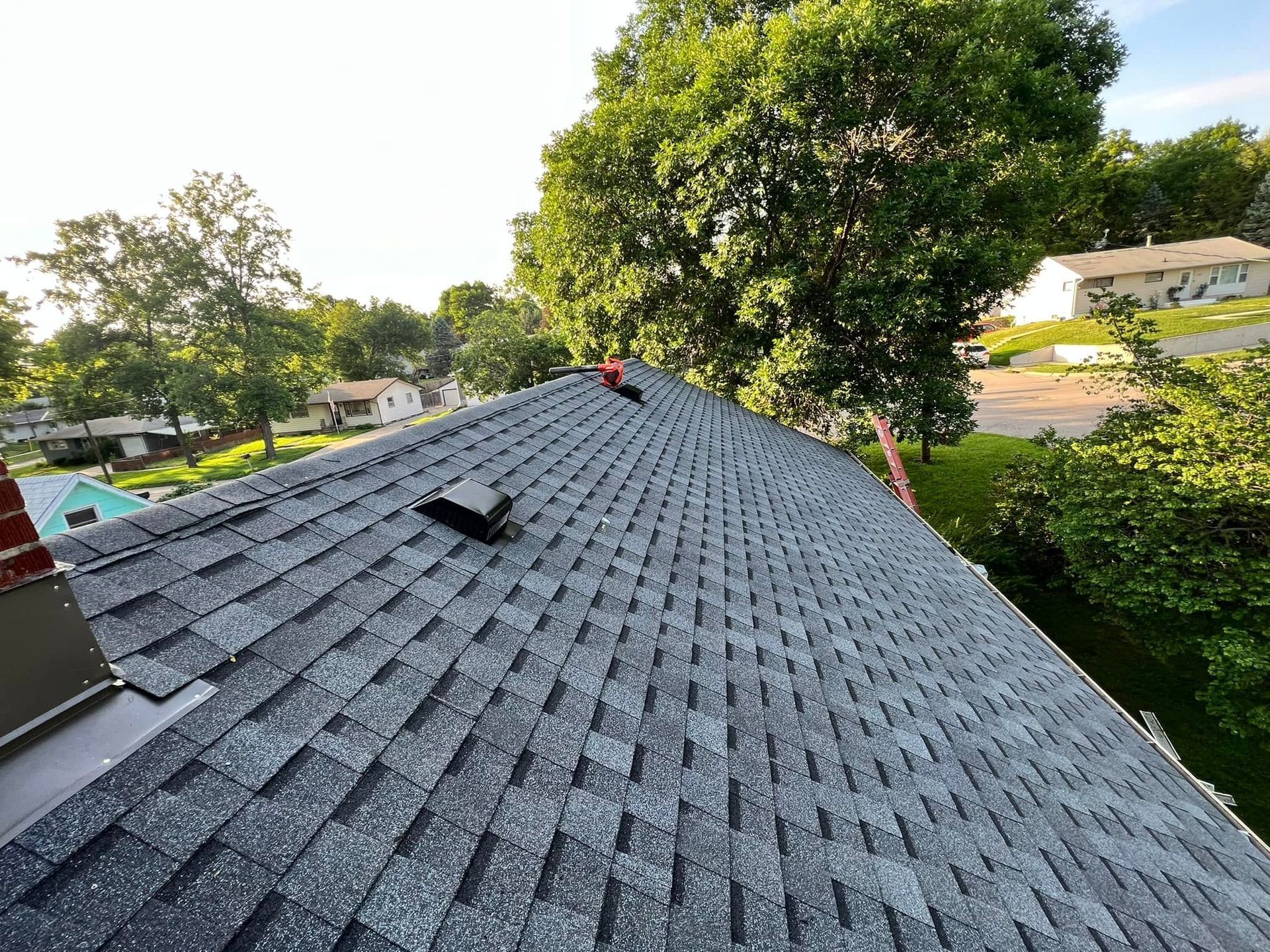Roof with dark gray shingles and two vents, viewed from above with trees and houses in the background