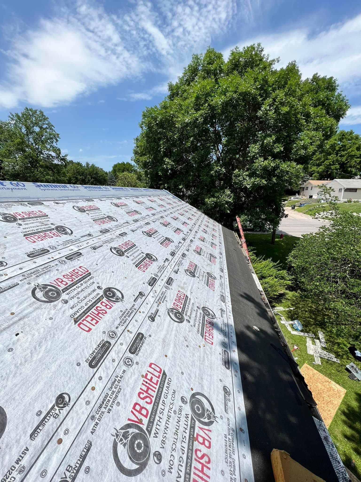 Roof underlayment beside leafy trees and a blue sky, viewed from a steep rooftop.