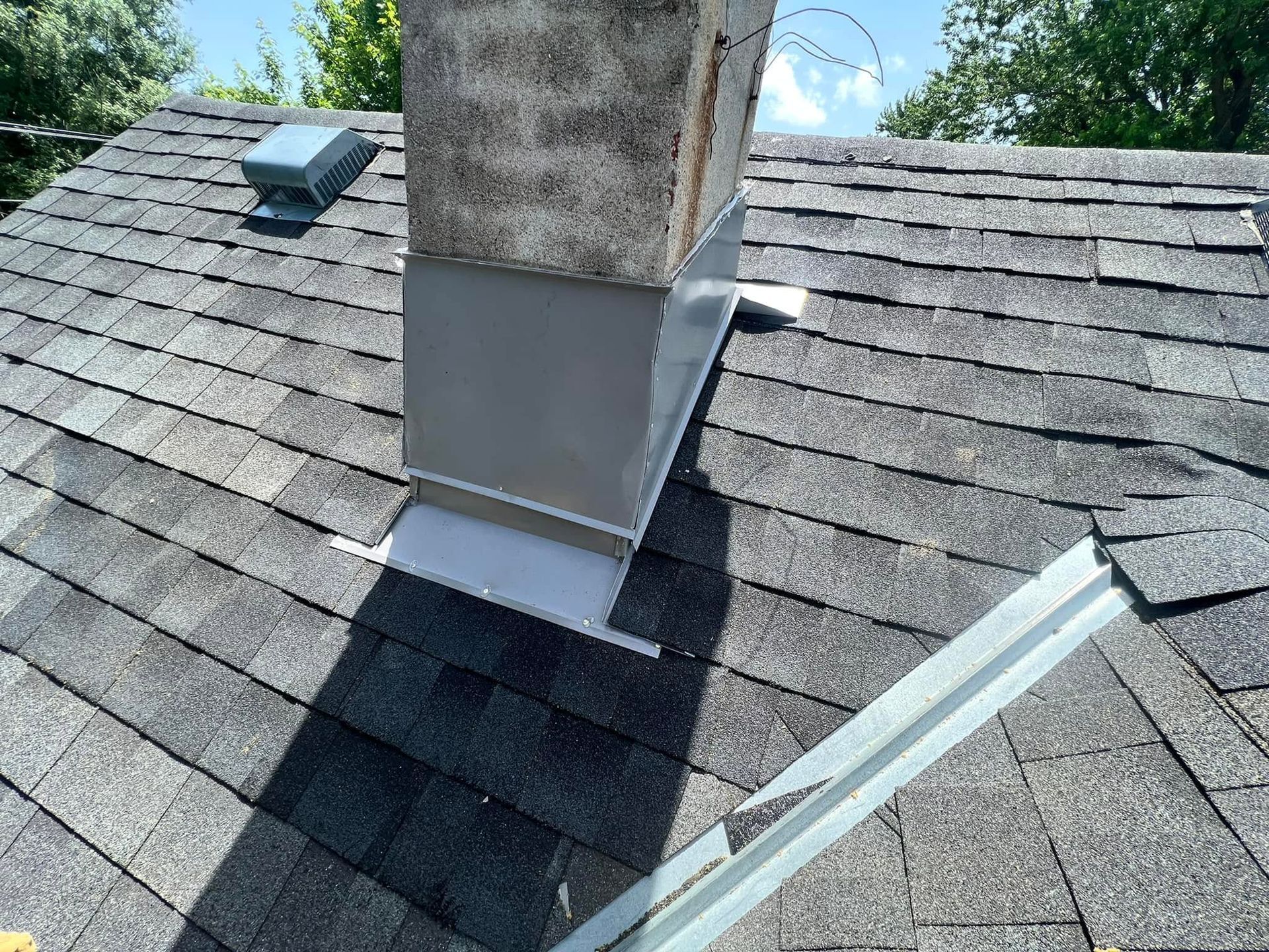 Rooftop chimney flashing repair on gray shingle roof, with metal vent and dormer skylight nearby