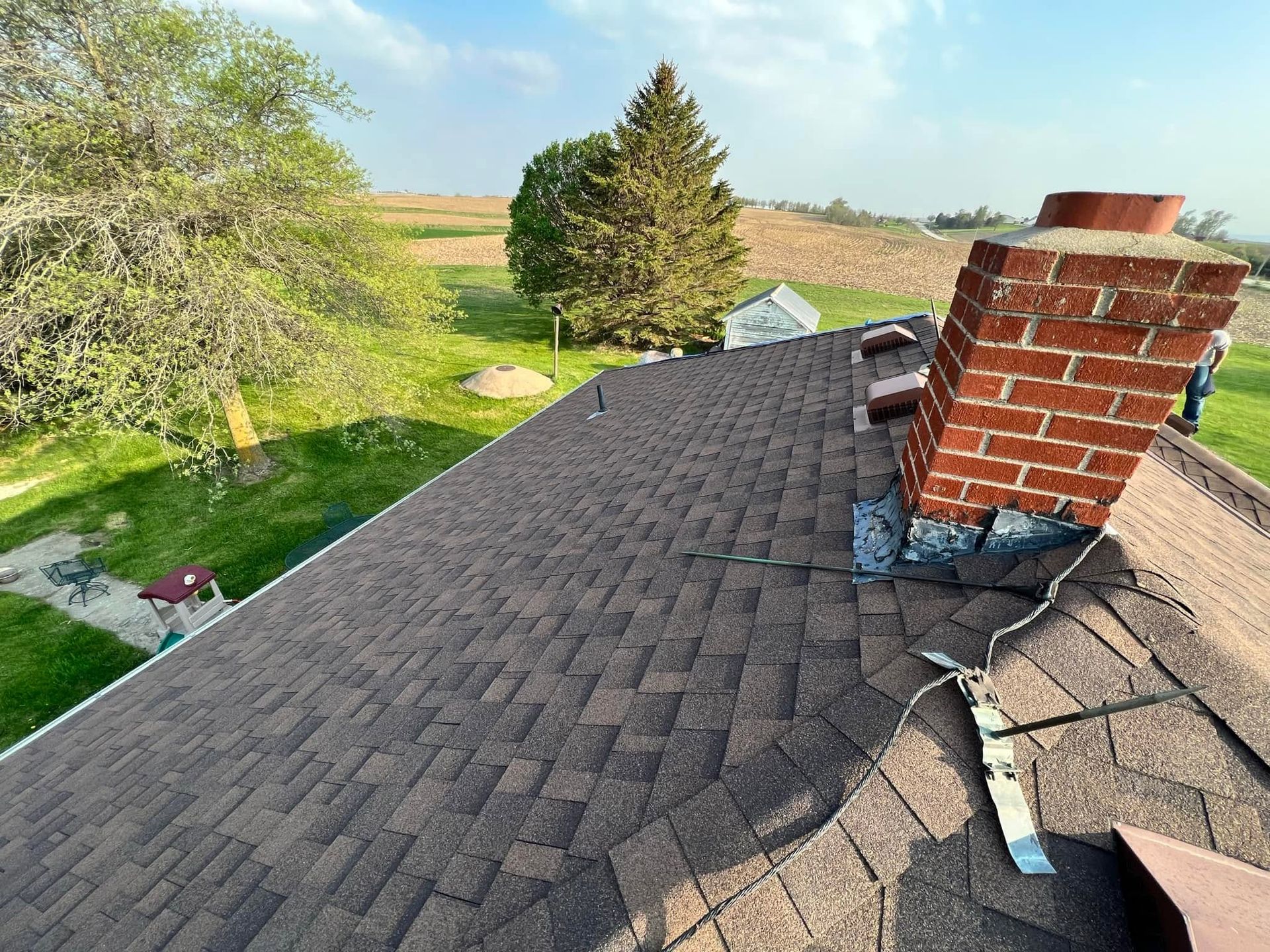 Roof shingles with a brick chimney and black cable beside a grassy yard and trees